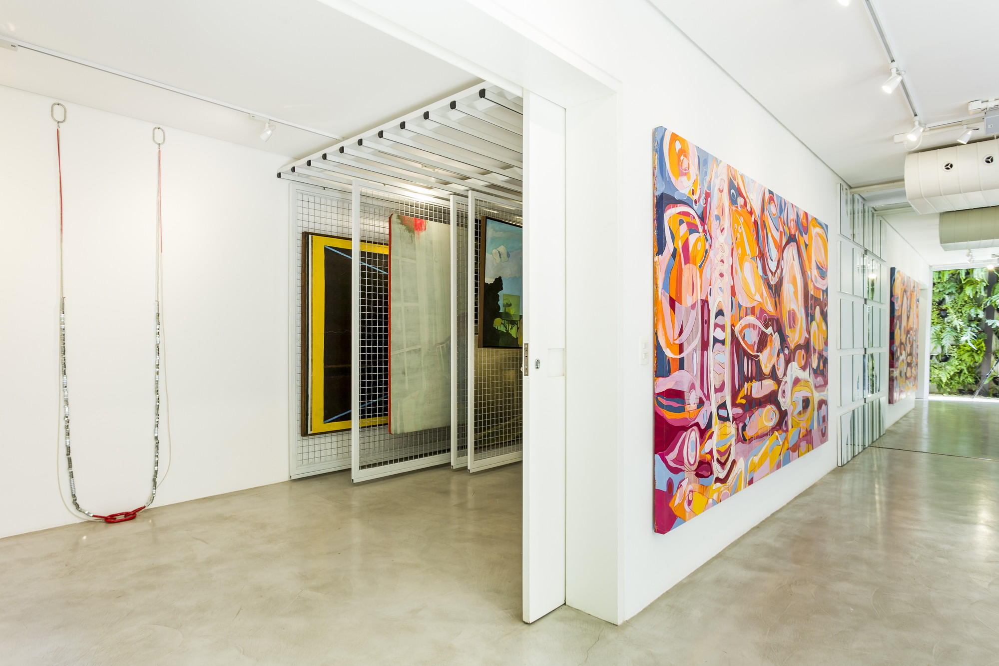 Corridor with a large abstract painting, a pulled-out mesh storage rack and a green planted courtyard glimpsed at the far end