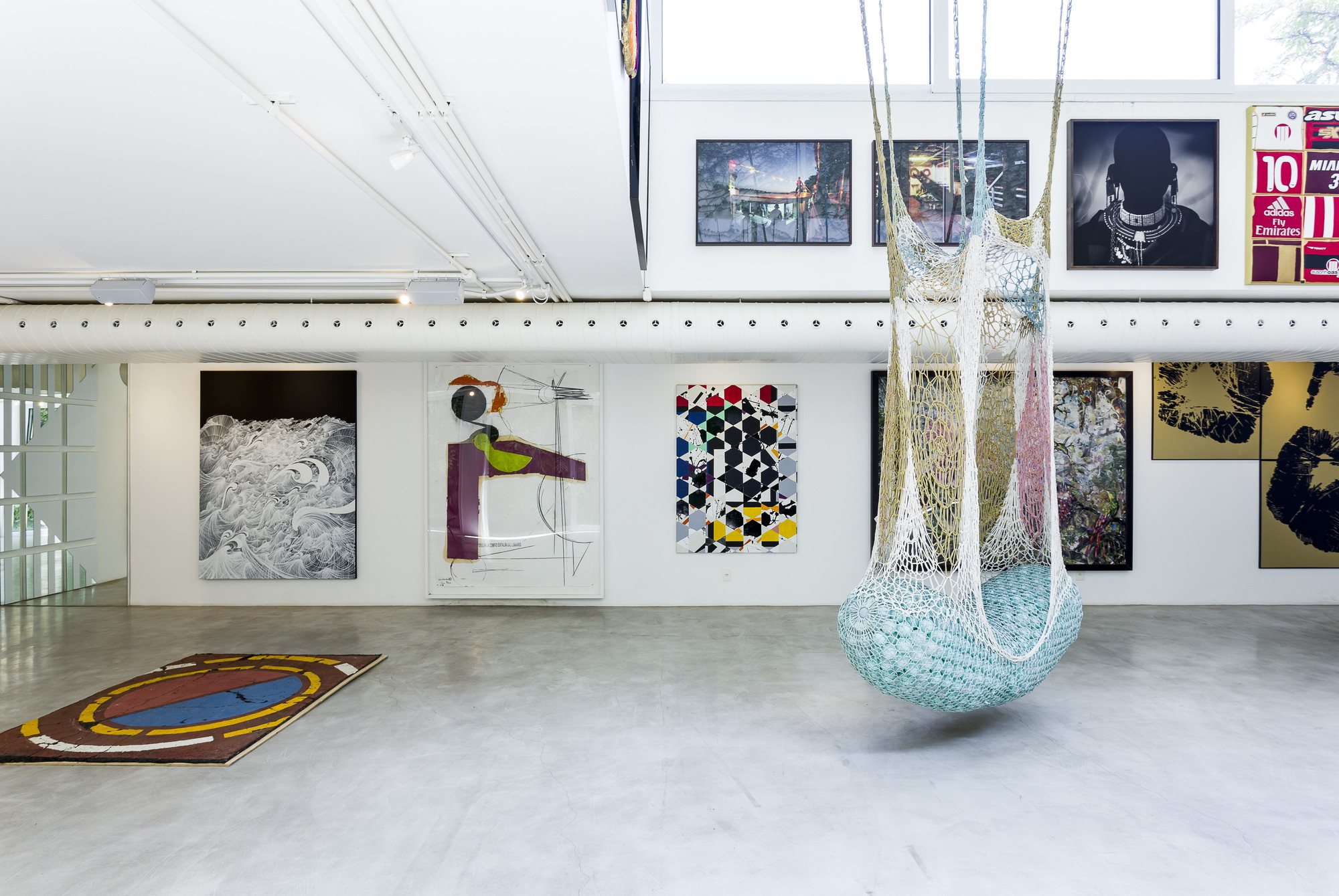 Gallery floor with a crocheted net hammock hanging in front of framed paintings and a perforated white duct under skylit clerestory windows