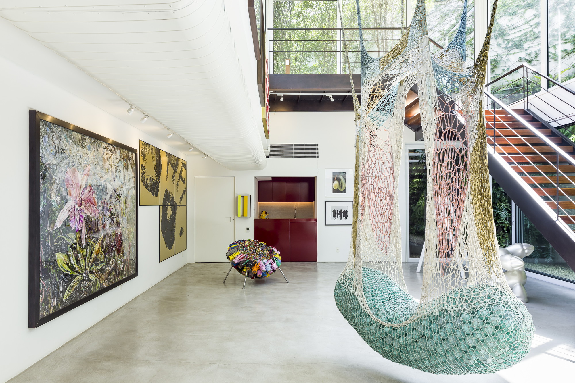 Double-height room with a hanging net seat, a large floral painting, a red cabinet and a timber stair, lit by trees outside