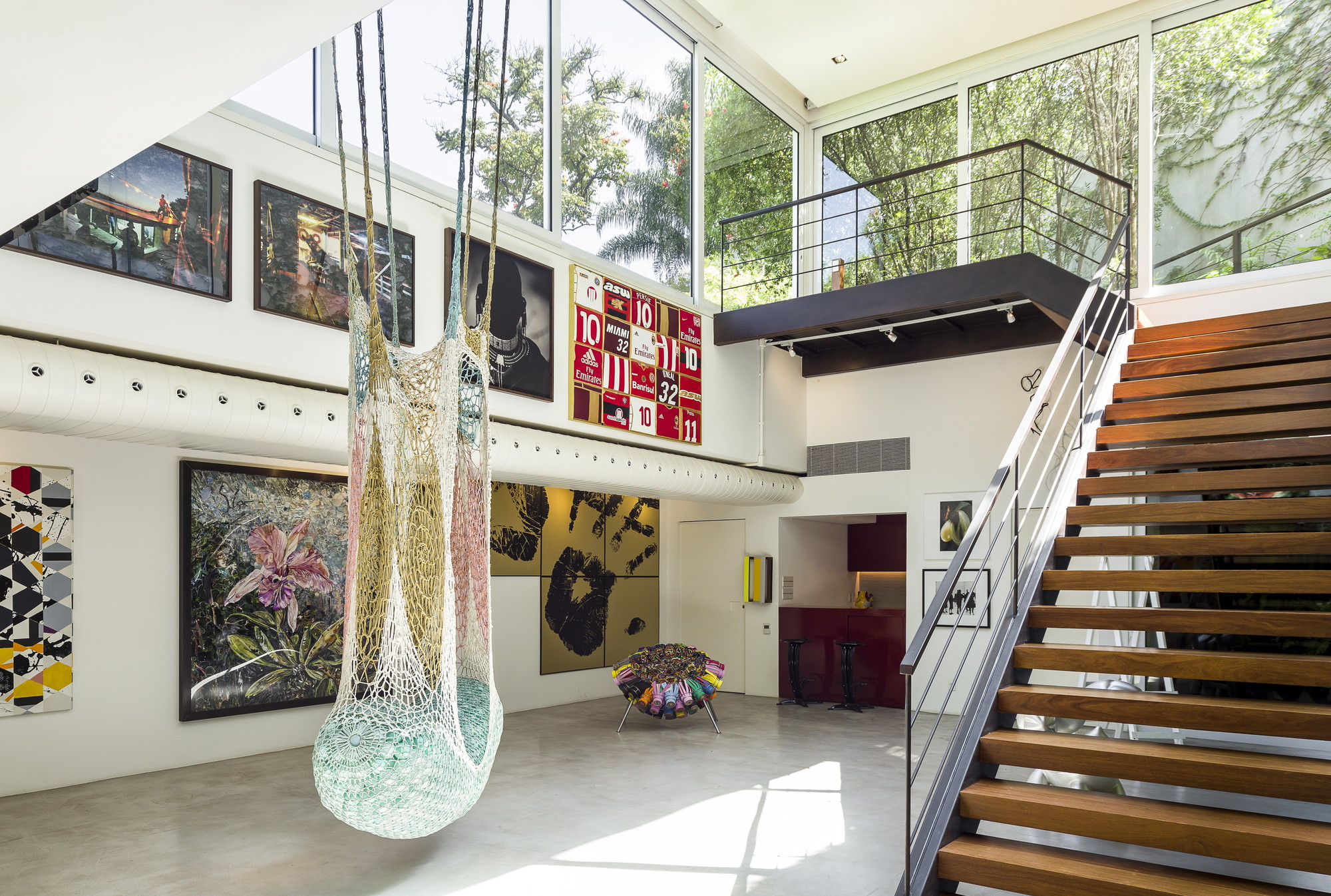 Open timber stair rising beside a double-height wall of art, with a net hammock hanging and sunlight patches on the concrete floor