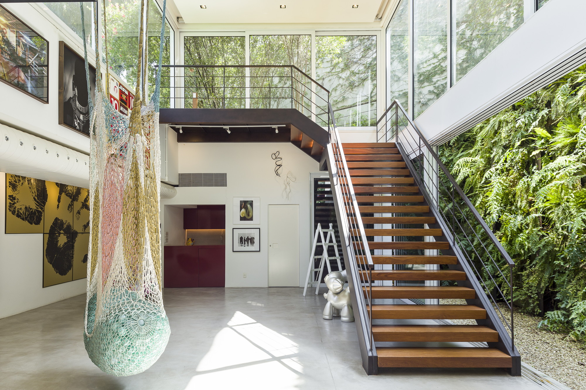 Steel and timber stair beside a fern-covered green wall, with a hanging net sculpture and window light falling across the concrete floor