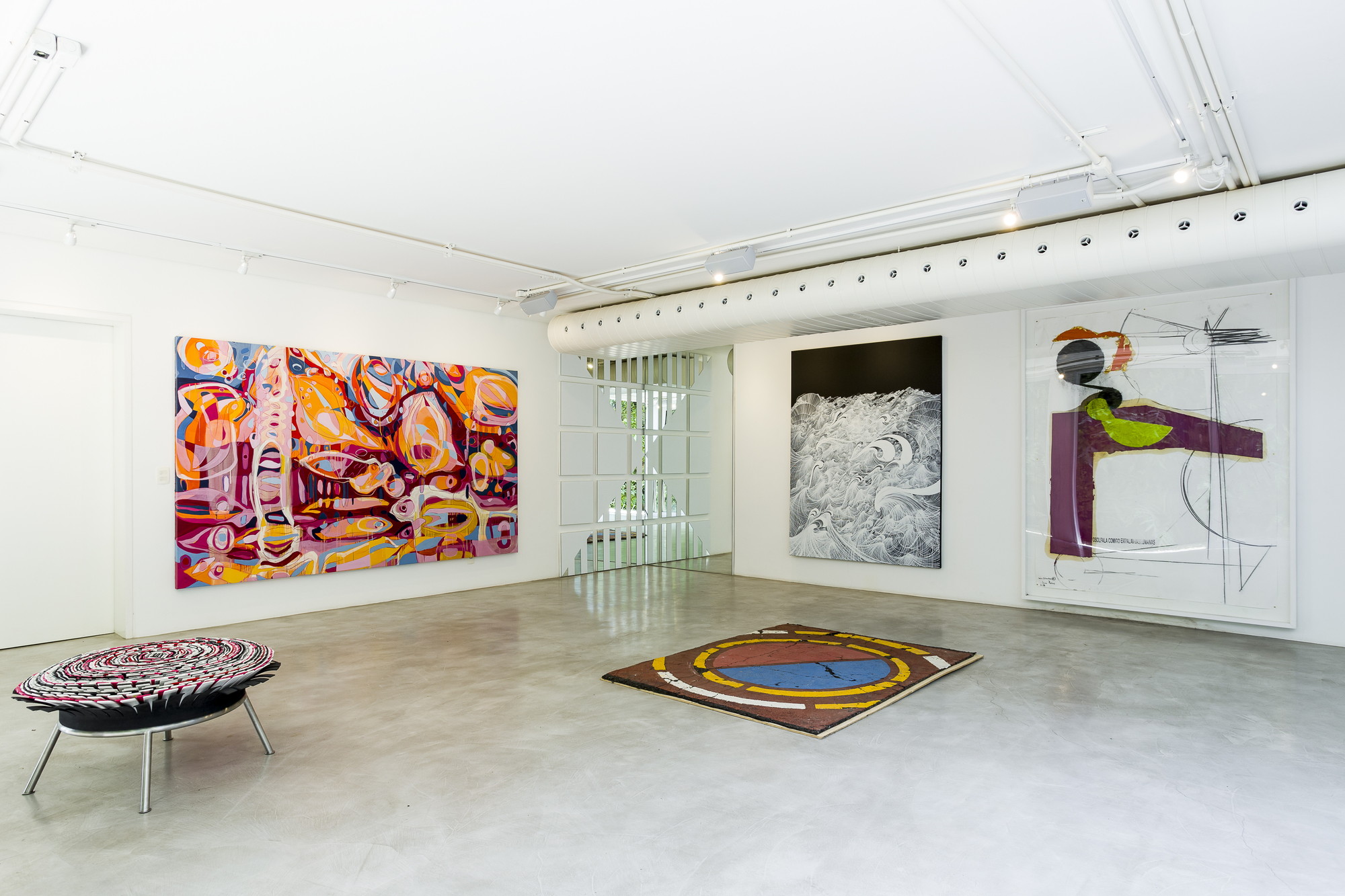 Gallery room with polished concrete floor, a round patterned rug, a woven stool and large paintings under a white ceiling duct