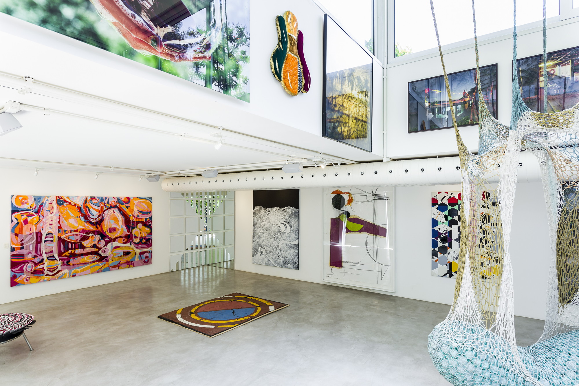 Double-height gallery with framed photographs on the upper wall and a crocheted net hammock hanging in the foreground in daylight