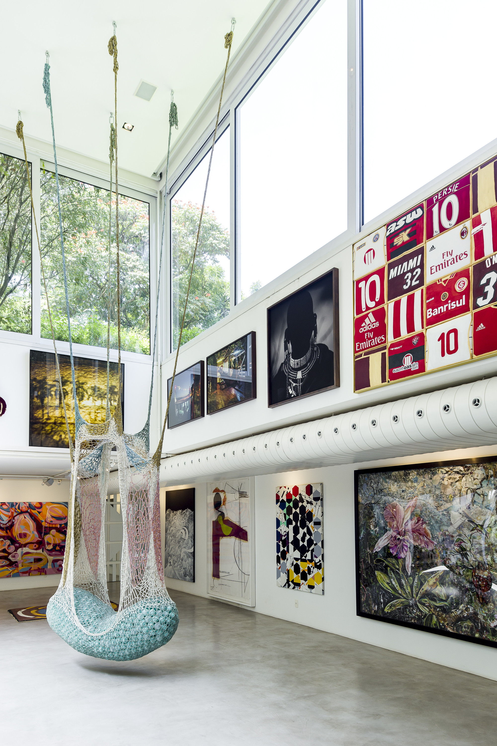 Tall gallery wall hung with framed football jerseys, photographs and a flower painting beside a suspended blue-green net chair