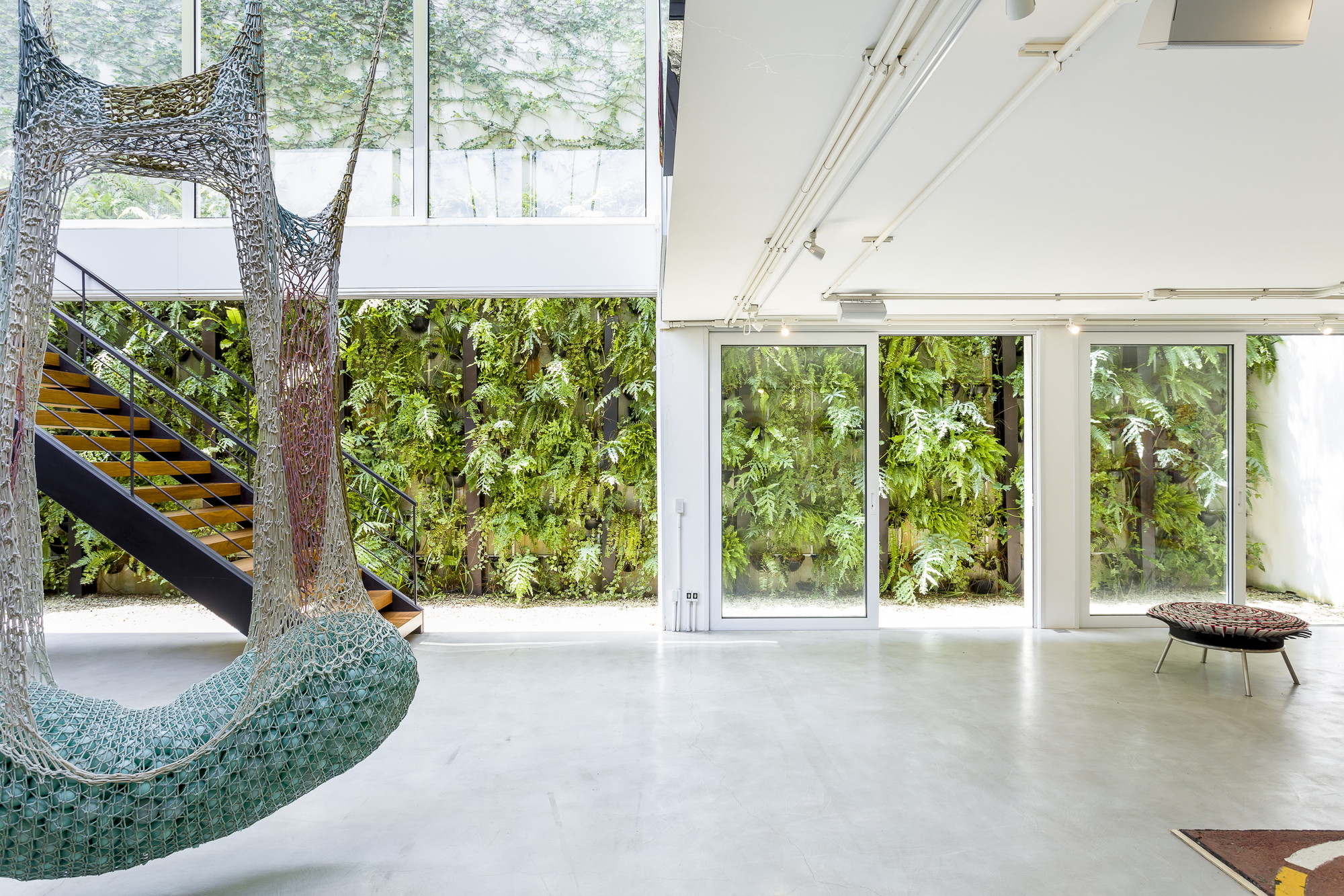 Steel and timber staircase beside a crocheted hanging seat, with a fern-covered green wall visible through tall glass doors