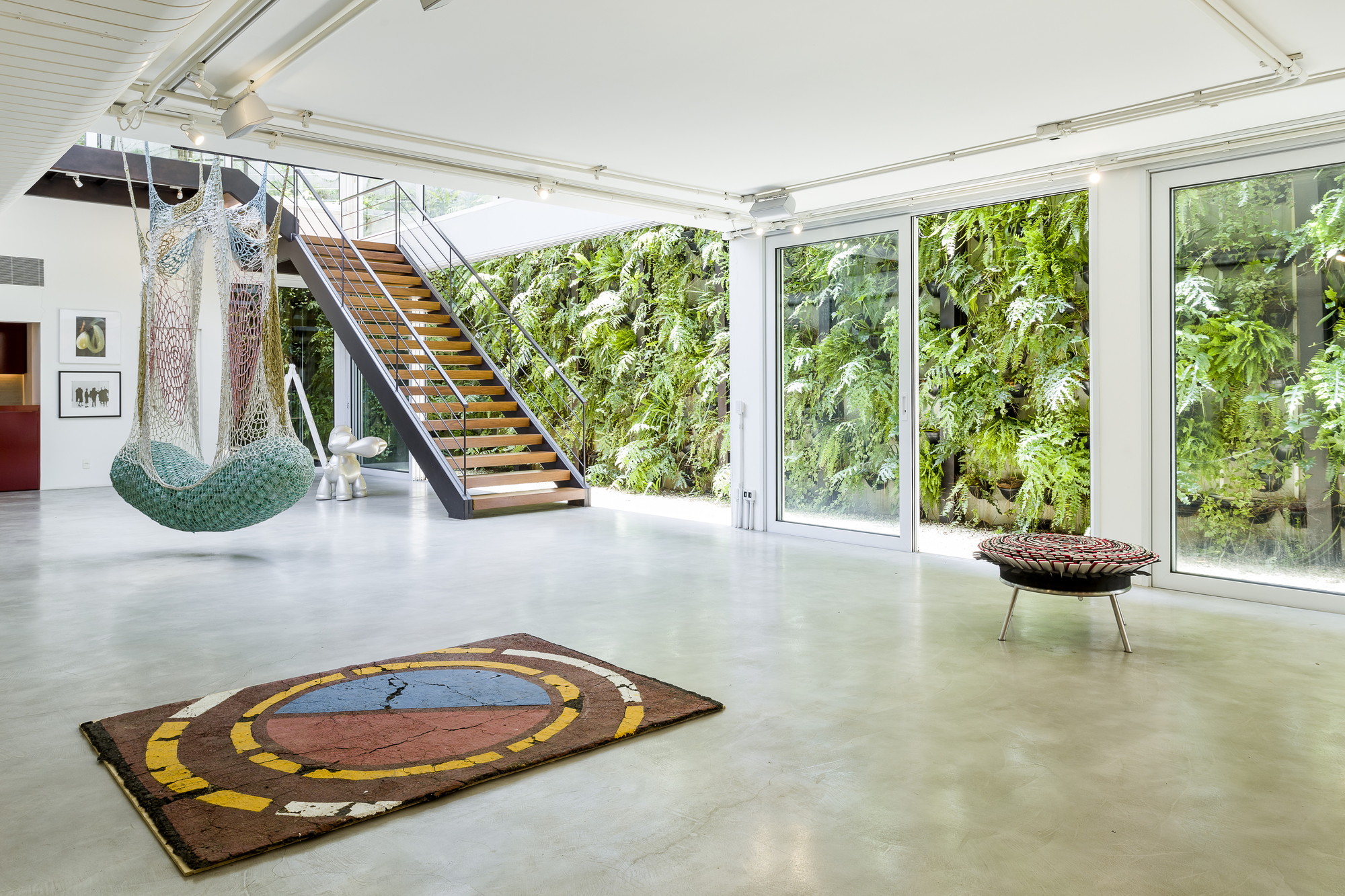 Open gallery floor with a circular rug, hanging net seat and timber stair facing a green wall of leafy plants