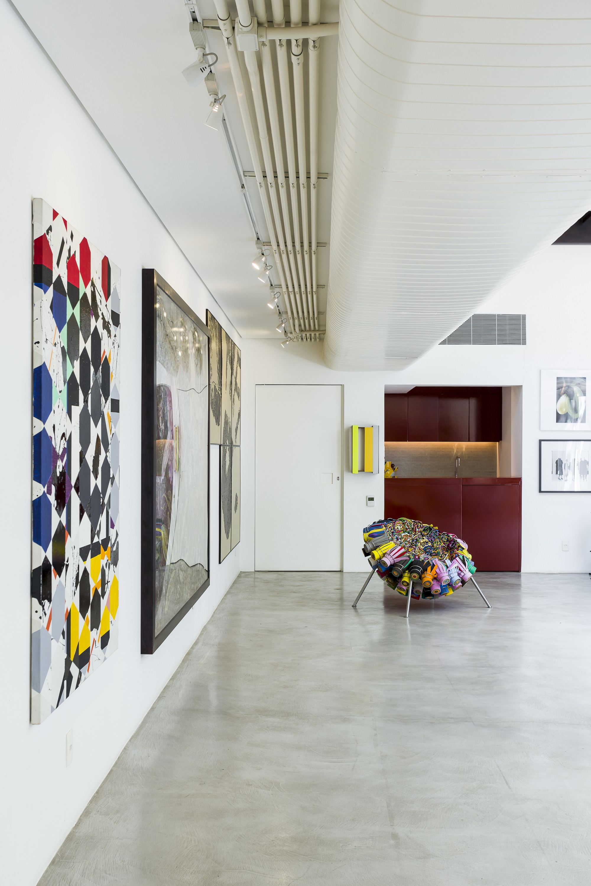 Gallery wall hung with colorful geometric paintings beside a sculptural patchwork chair and a burgundy cabinet niche under a curved white ceiling duct