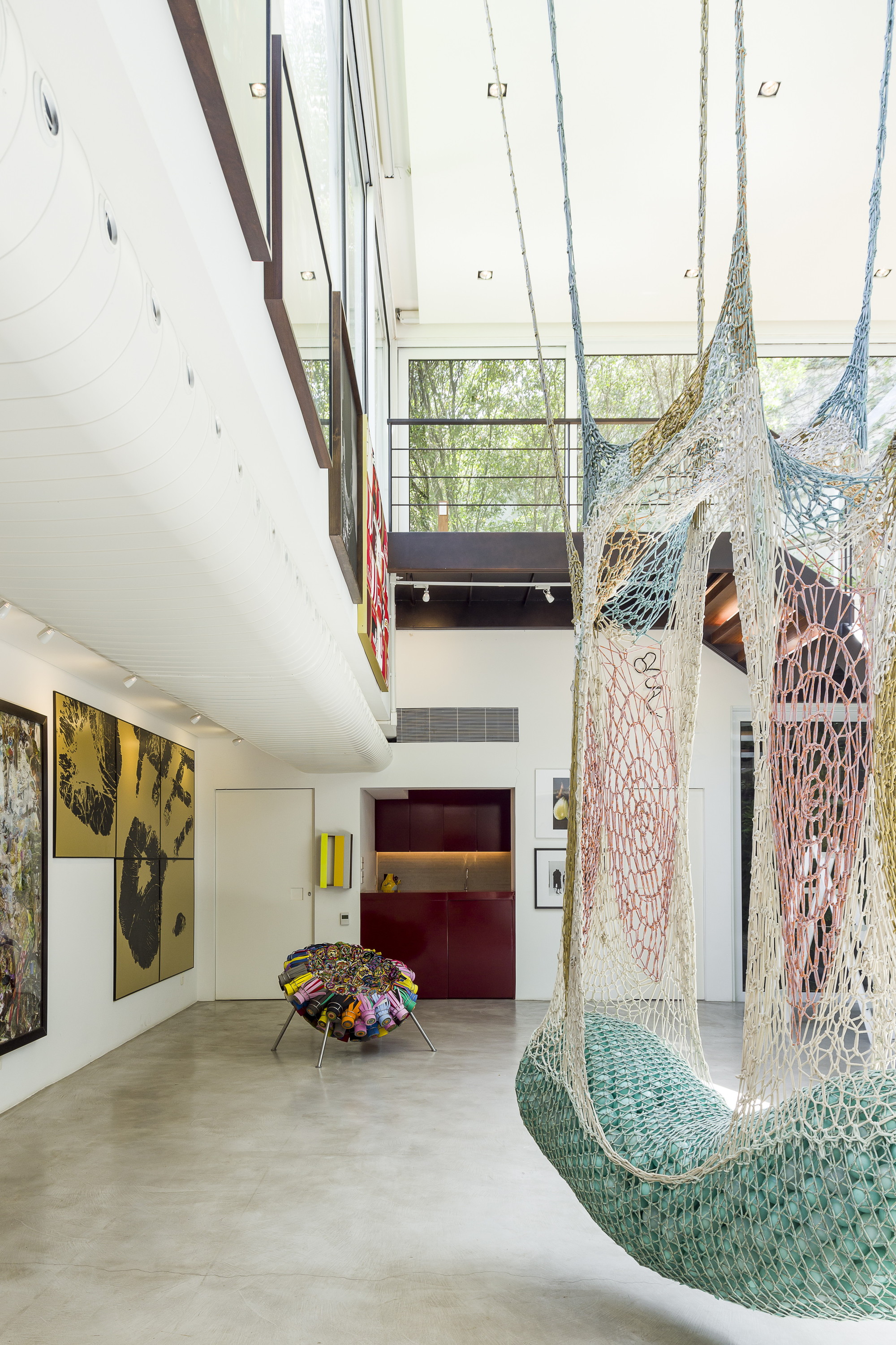 Double-height gallery with a hanging crocheted net sculpture, a mezzanine walkway and trees visible through glazing in daylight