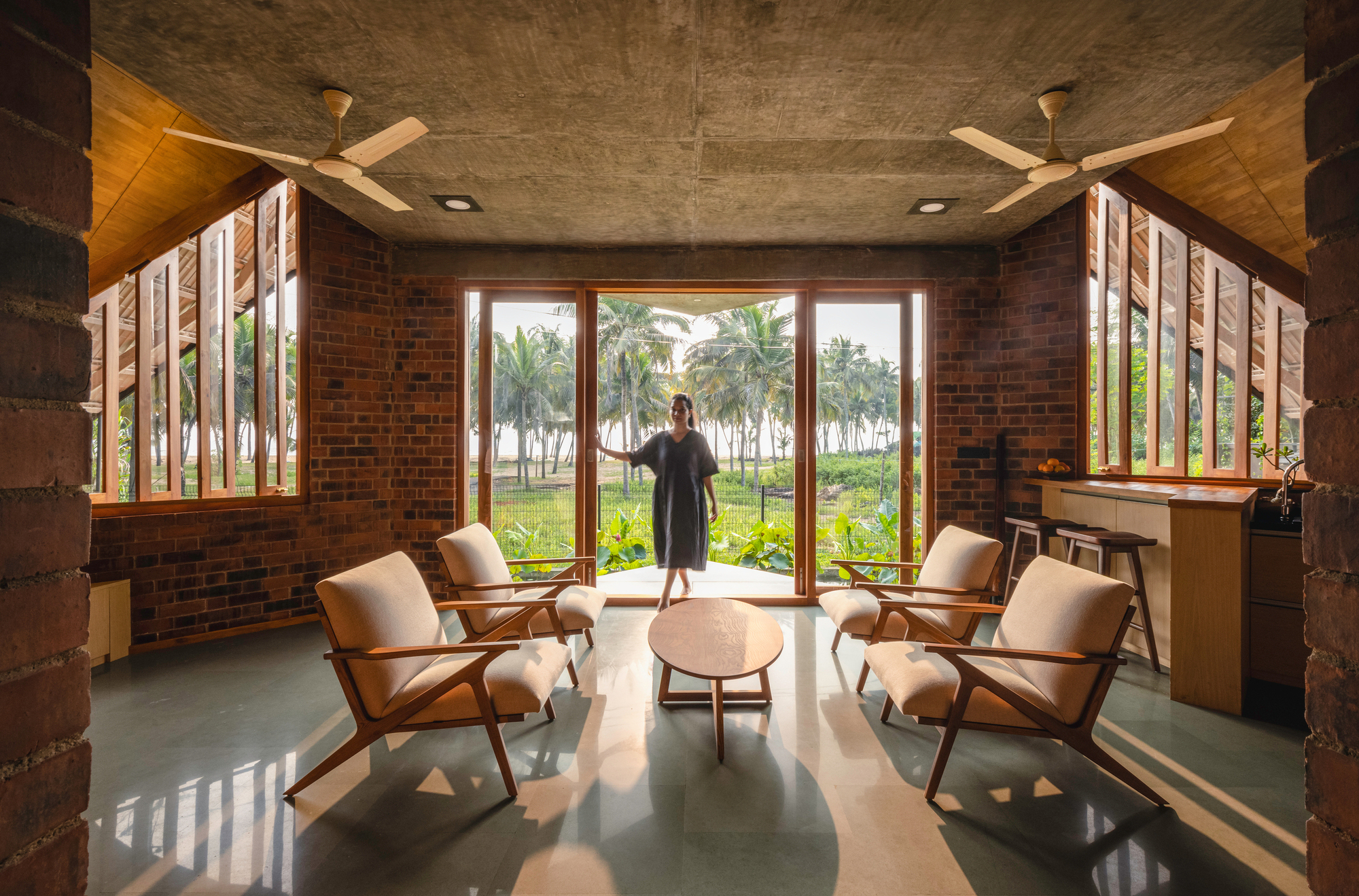 Living room with red brick walls and tall slatted windows framing a palm grove with a figure at the glass door