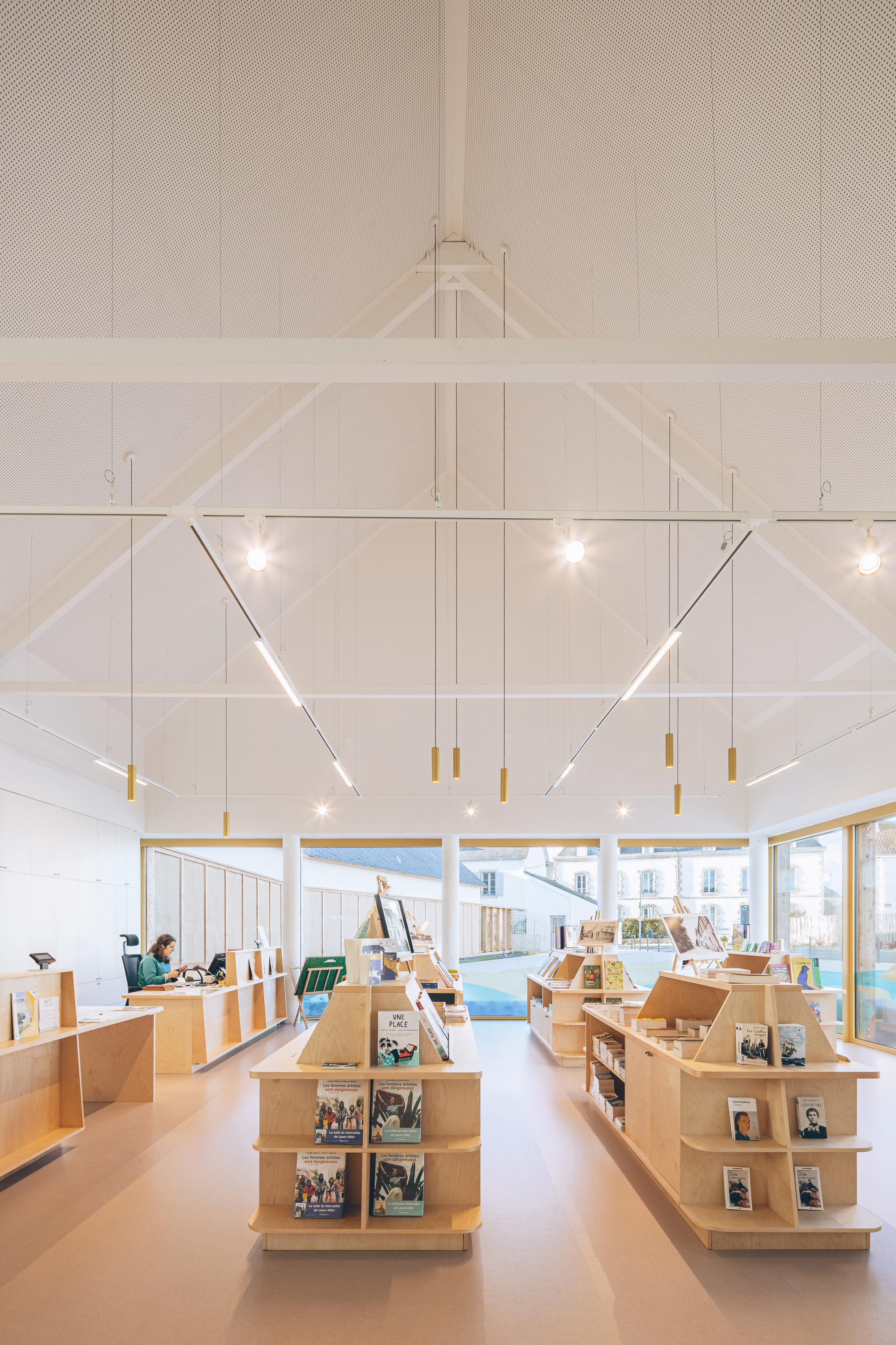 Interior library space with white paneled walls, exposed timber roof trusses and rows of blonde wood shelving units
