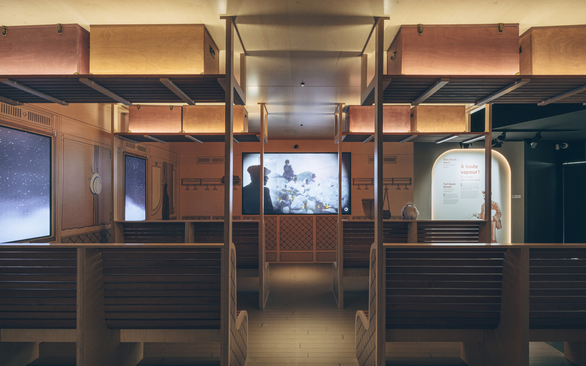Exhibition gallery with timber-clad benches, overhead mezzanine structure and illuminated video screens at night