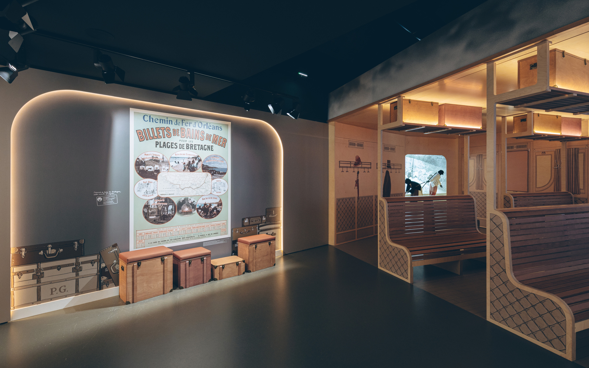 Museum gallery featuring a backlit poster wall, historic luggage and wooden railway carriage pews under black ceiling