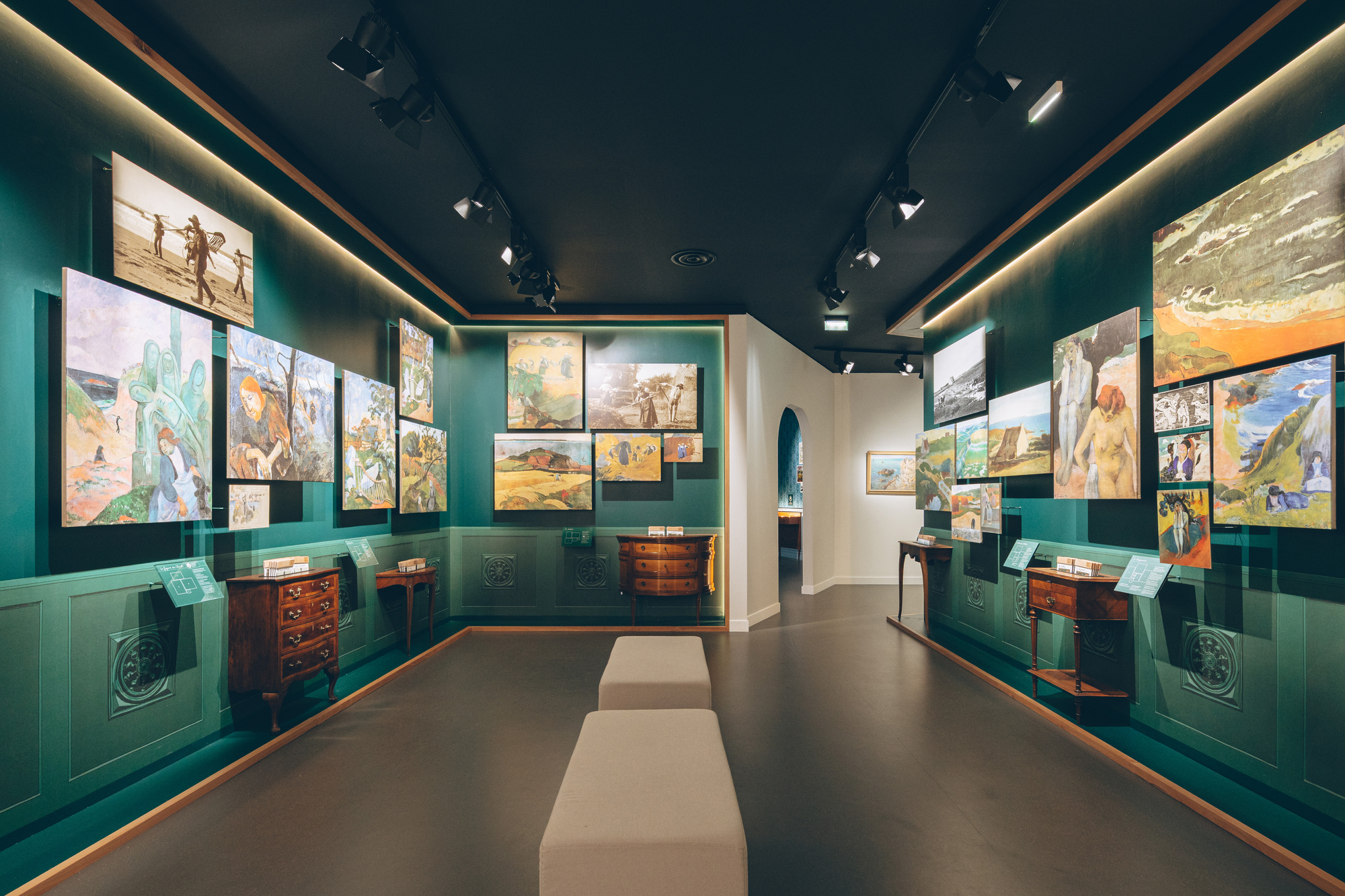 Gallery corridor with teal paneled walls, framed paintings hung in rows and central upholstered bench seating