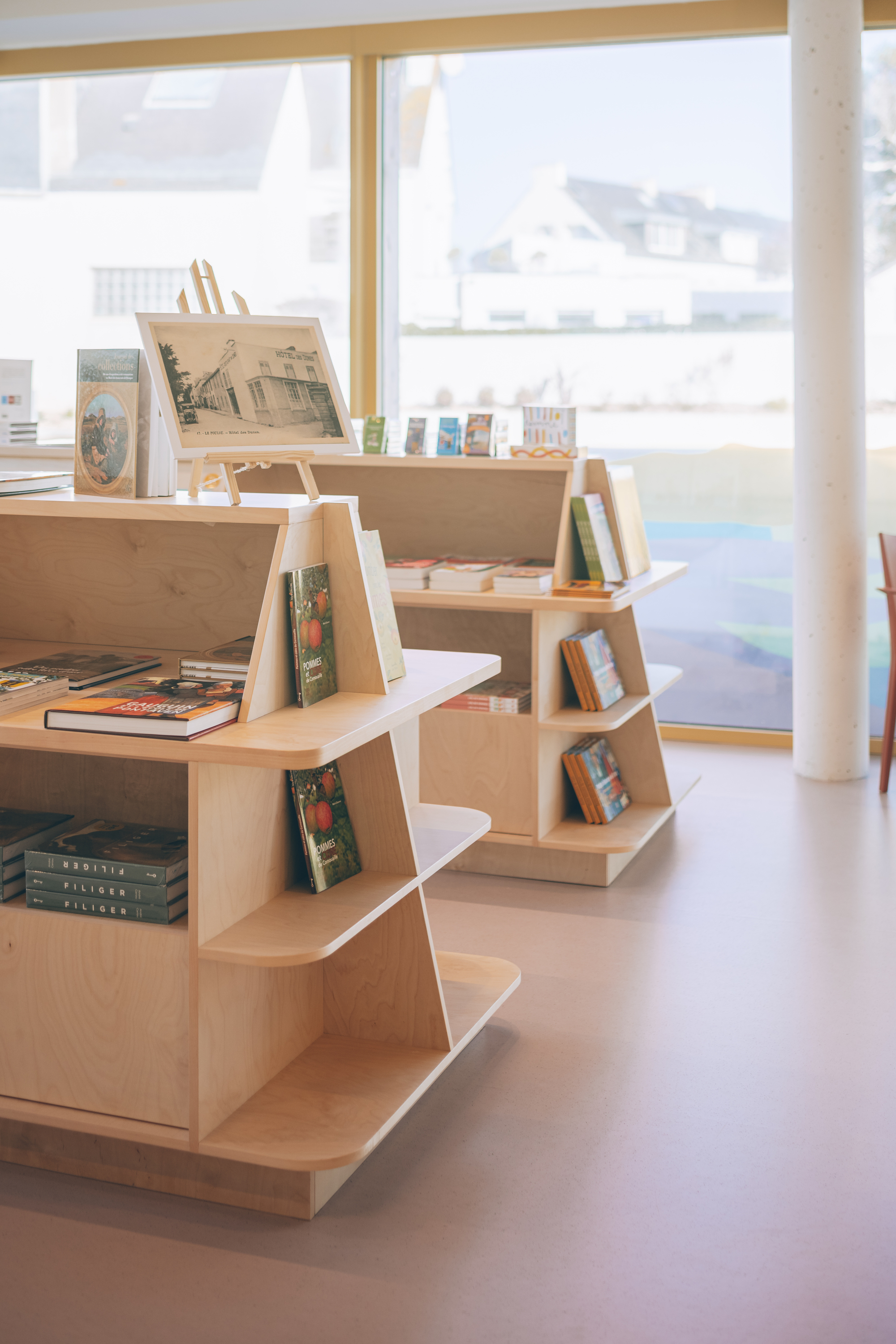 Stepped timber display shelves holding books and objects beneath full-height glazing and a concrete column