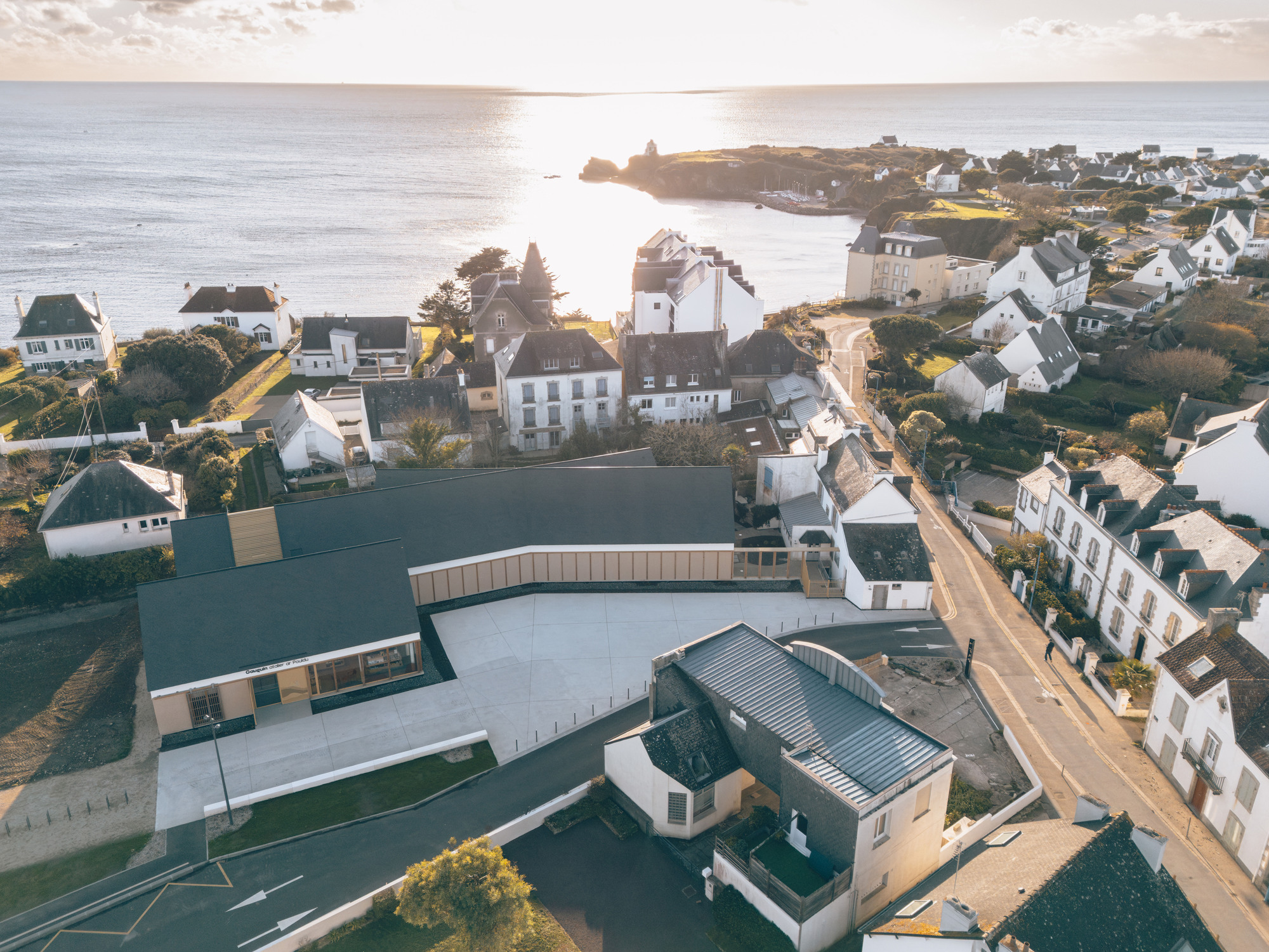 Aerial view of angled zinc roofs arranged around a central courtyard near a coastal village at golden hour