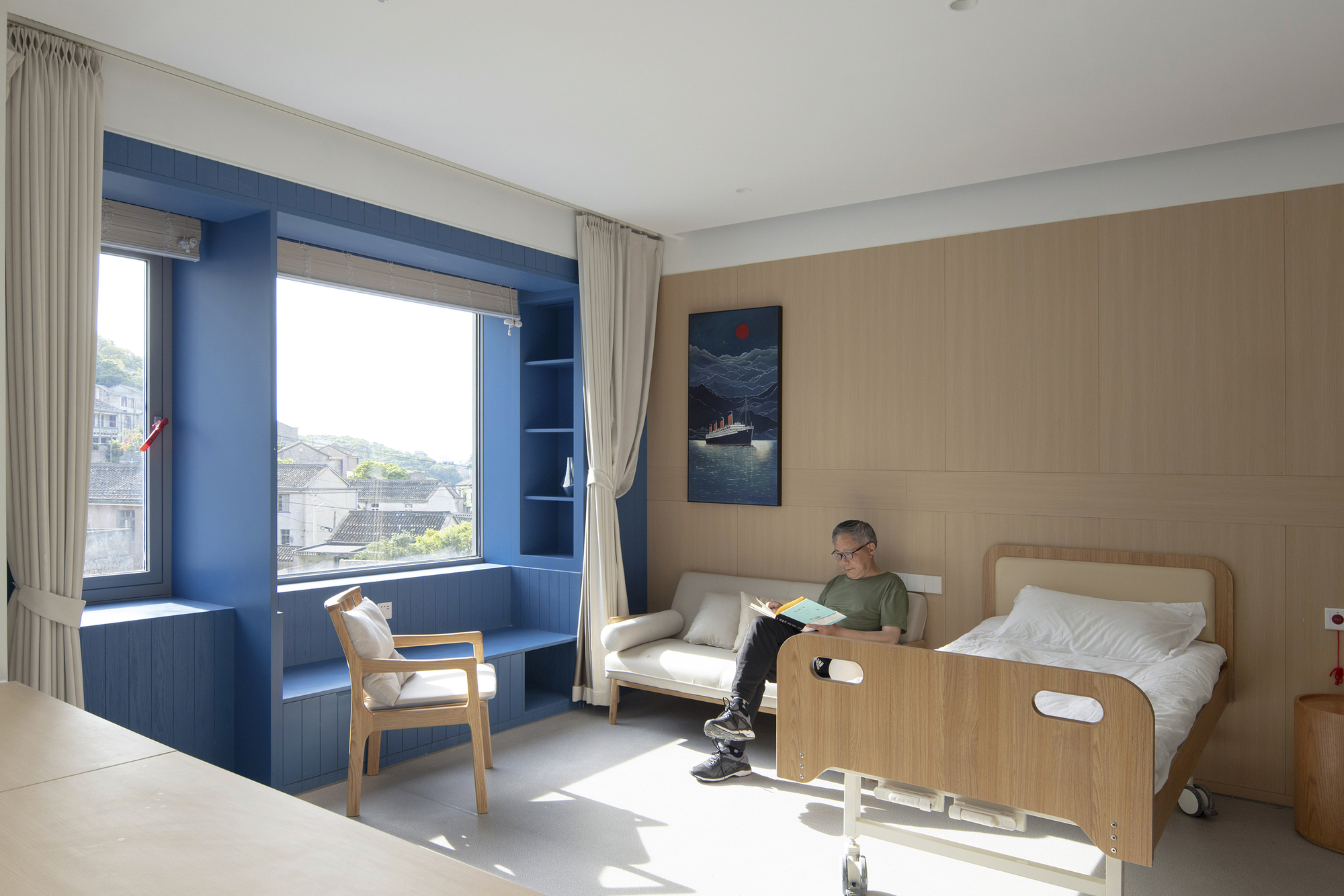 Patient room with timber wall panels, blue window alcove, and a person reading beside the bed