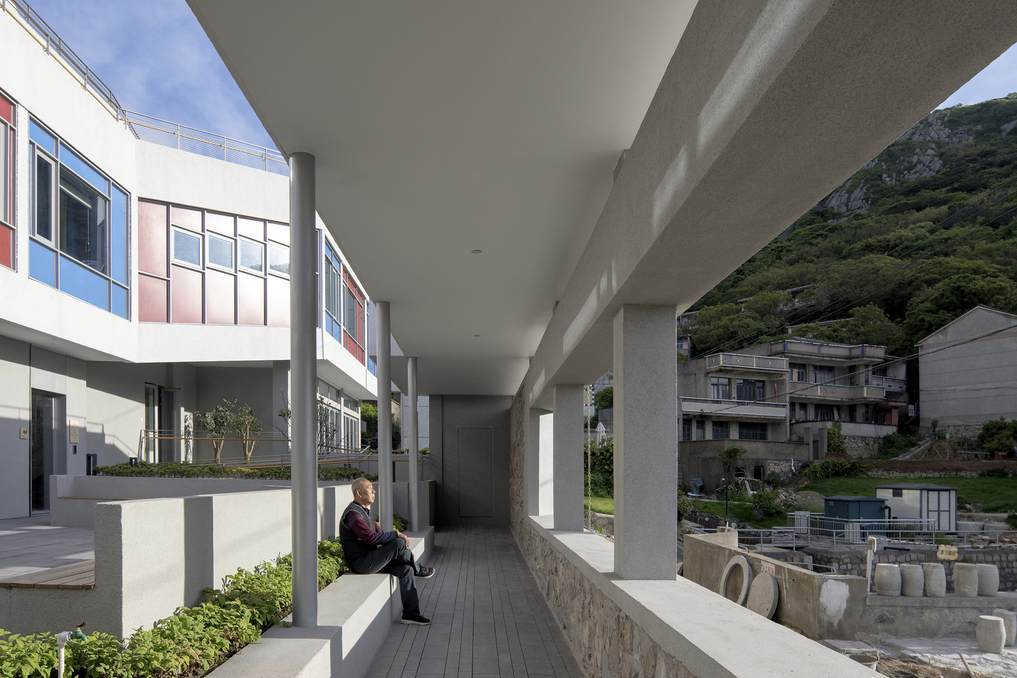 Covered corridor with concrete columns and planted beds where a person sits reading