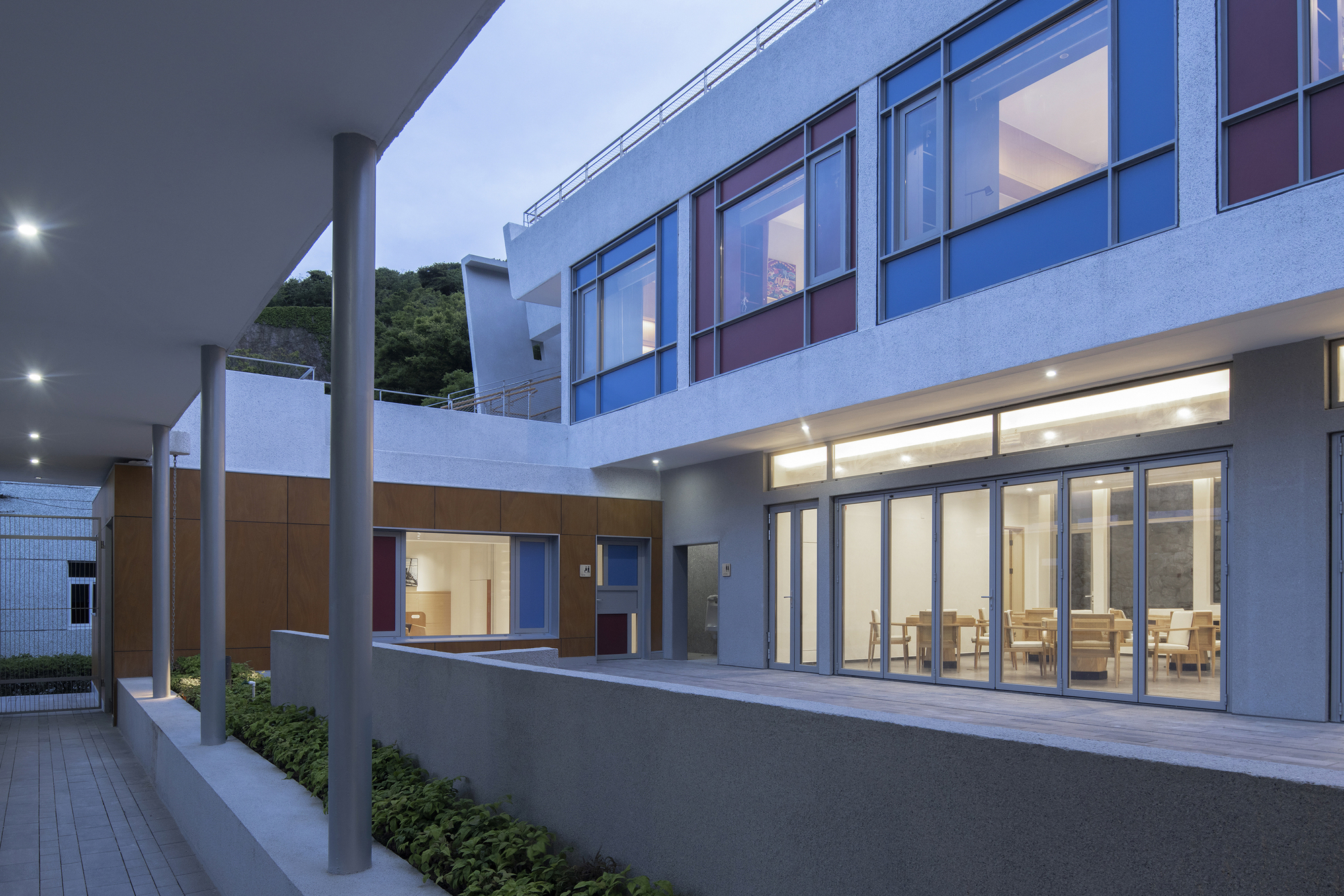 Courtyard view at dusk with ground-floor dining hall glazing and cantilevered columns along the entry drive