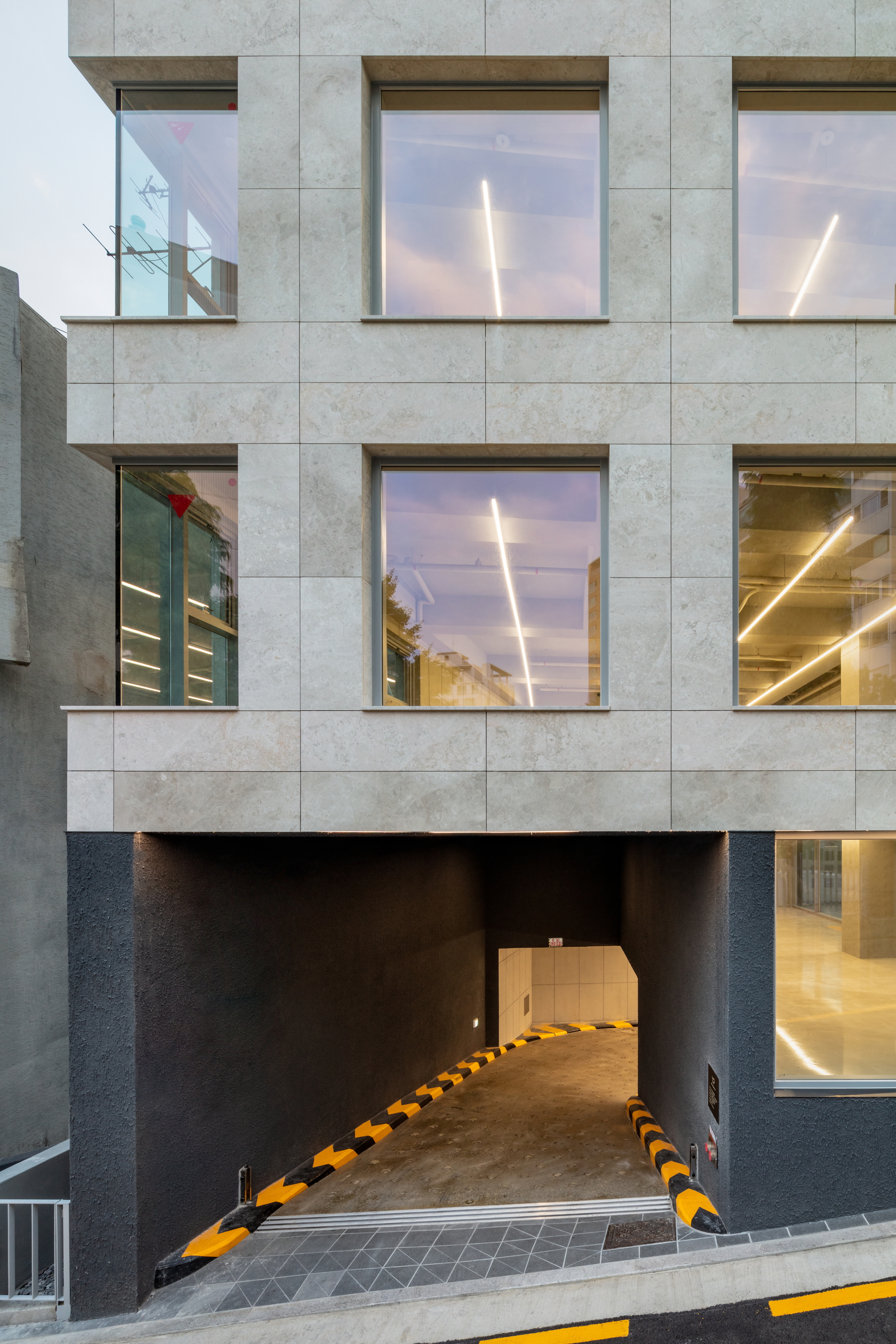 Ground level entrance showing a dark vehicular ramp beneath stone-clad upper floors at dusk