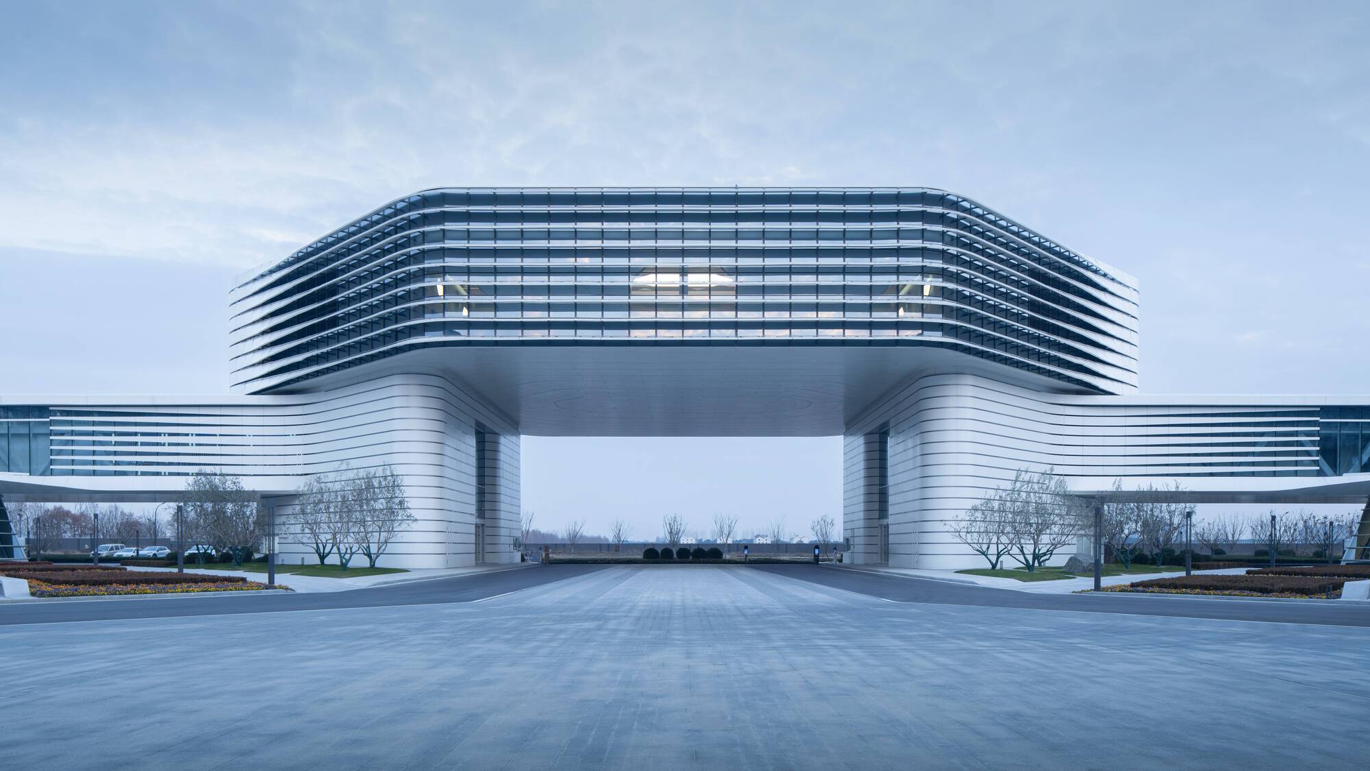 Frontal view of the elevated horizontal volume supported by curved ribbed columns forming a plaza gateway on an overcast day