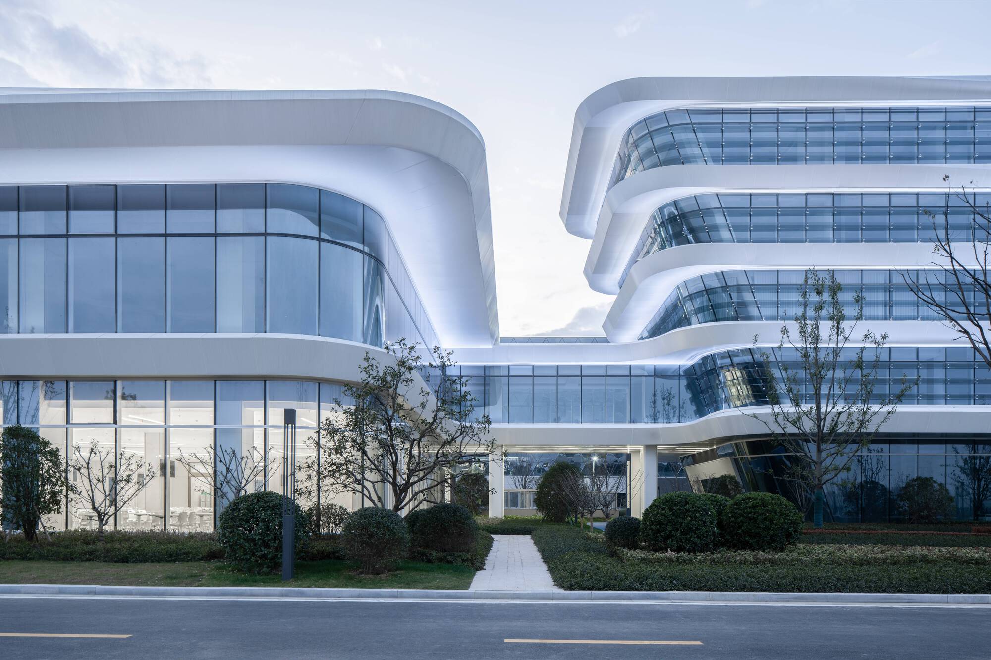Courtyard view through rounded glass facades with connecting bridge above manicured hedge plantings