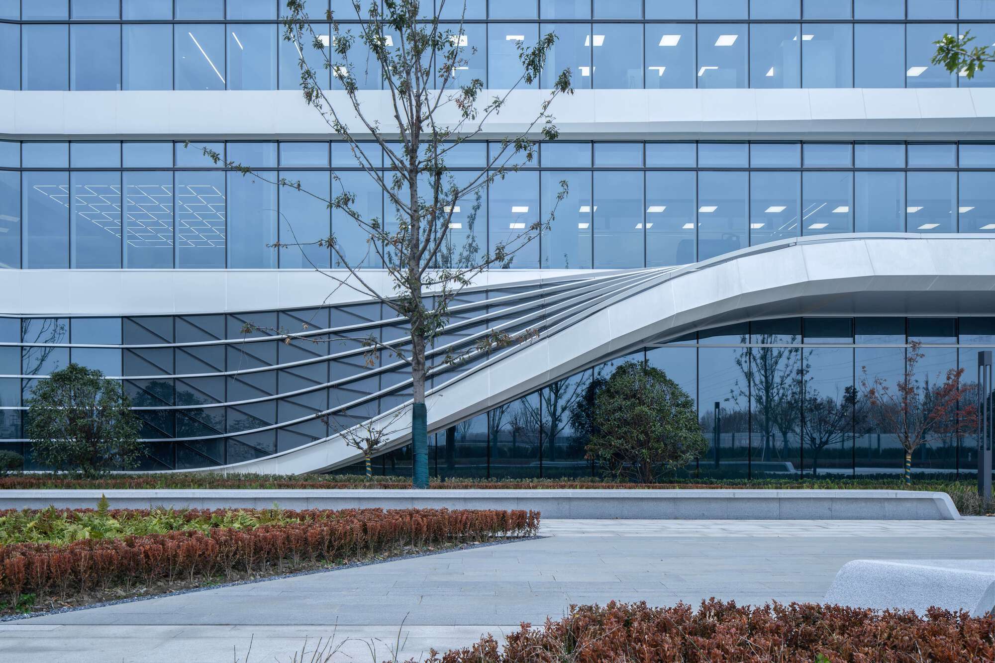 Curved pedestrian bridge spanning a landscaped courtyard with planted beds and young trees