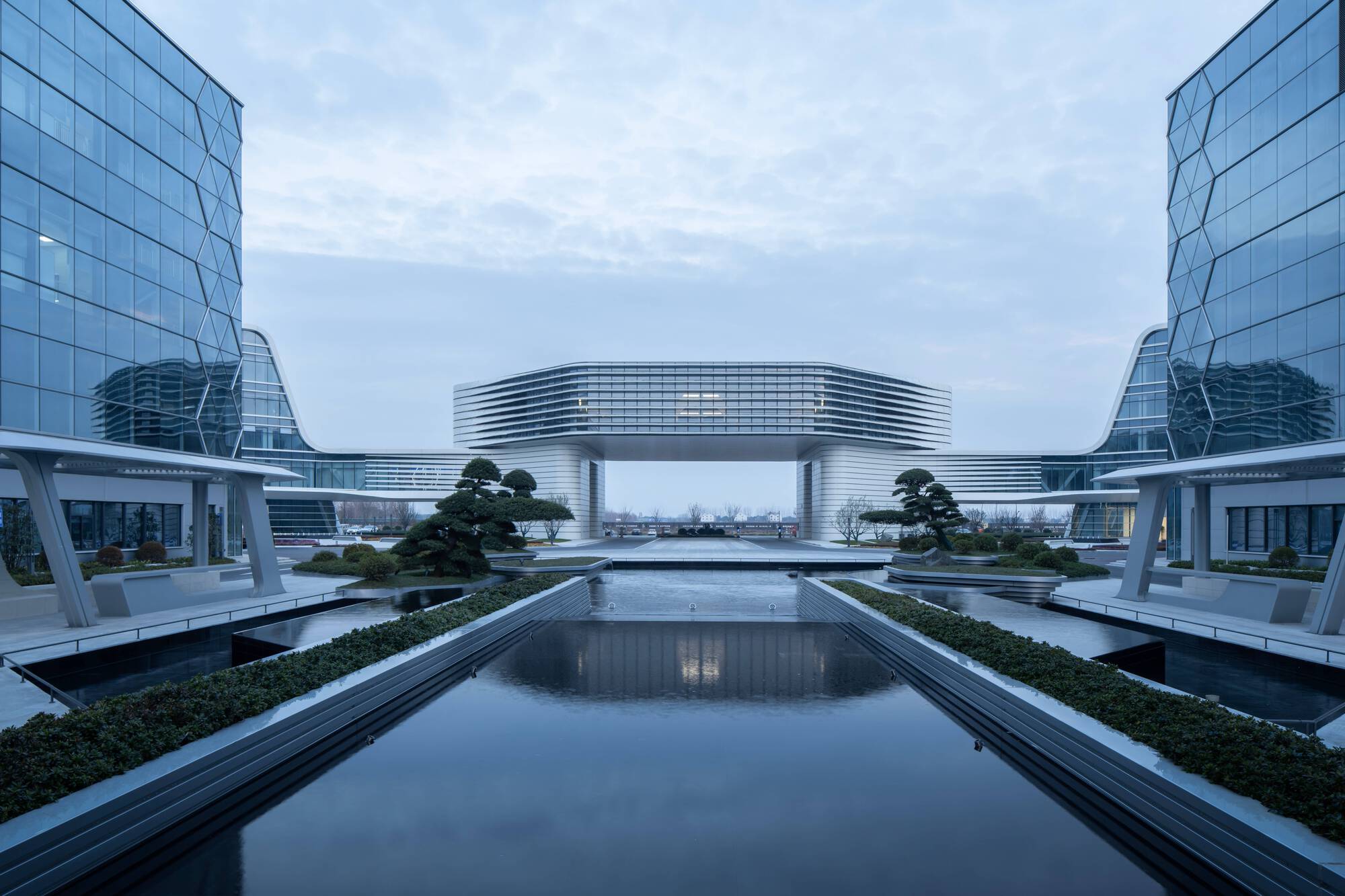 Symmetrical water feature with bridge structure connecting glass towers under an overcast sky at twilight