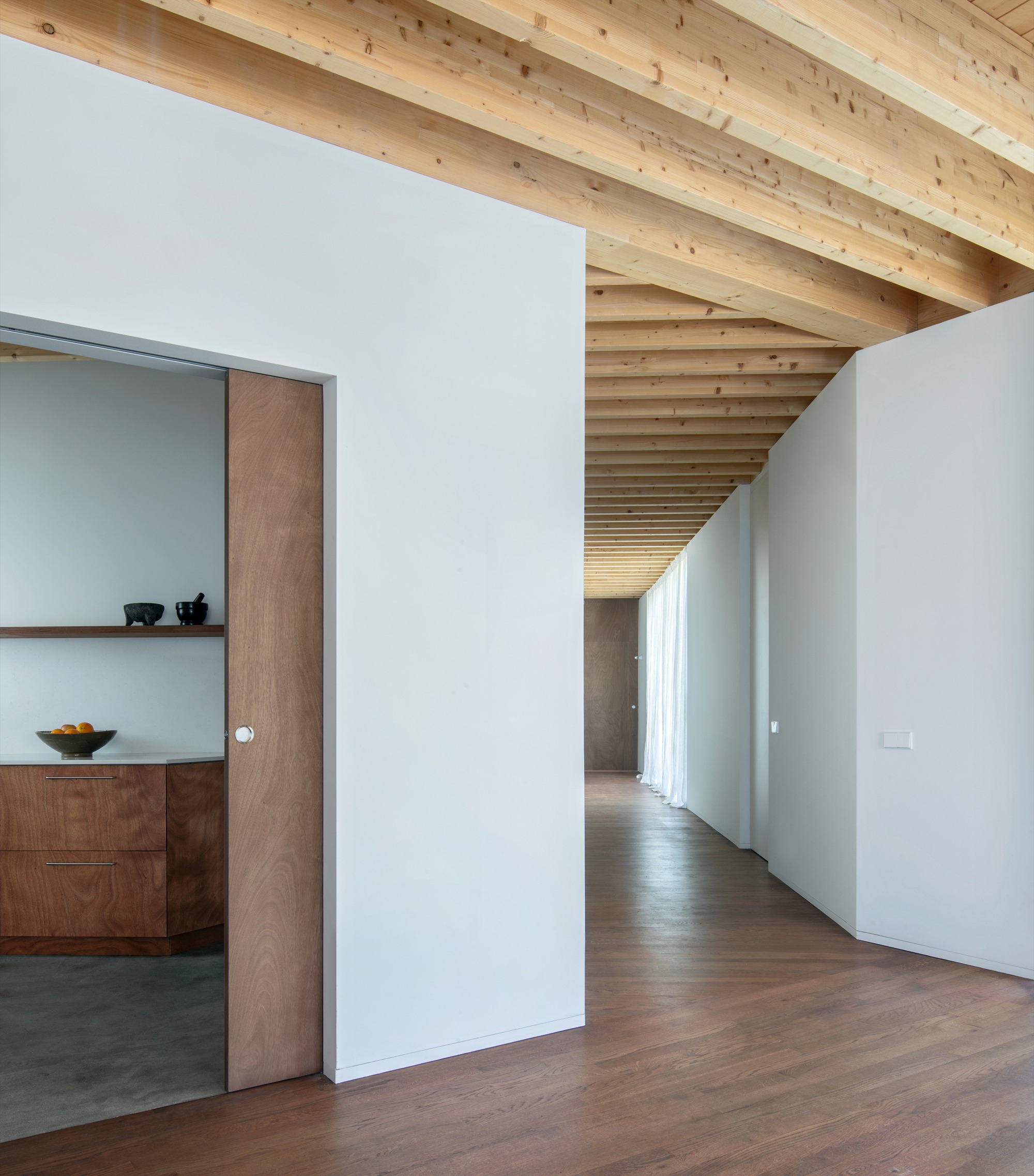 Interior hallway with exposed timber joists and white walls opening to kitchen with walnut cabinetry