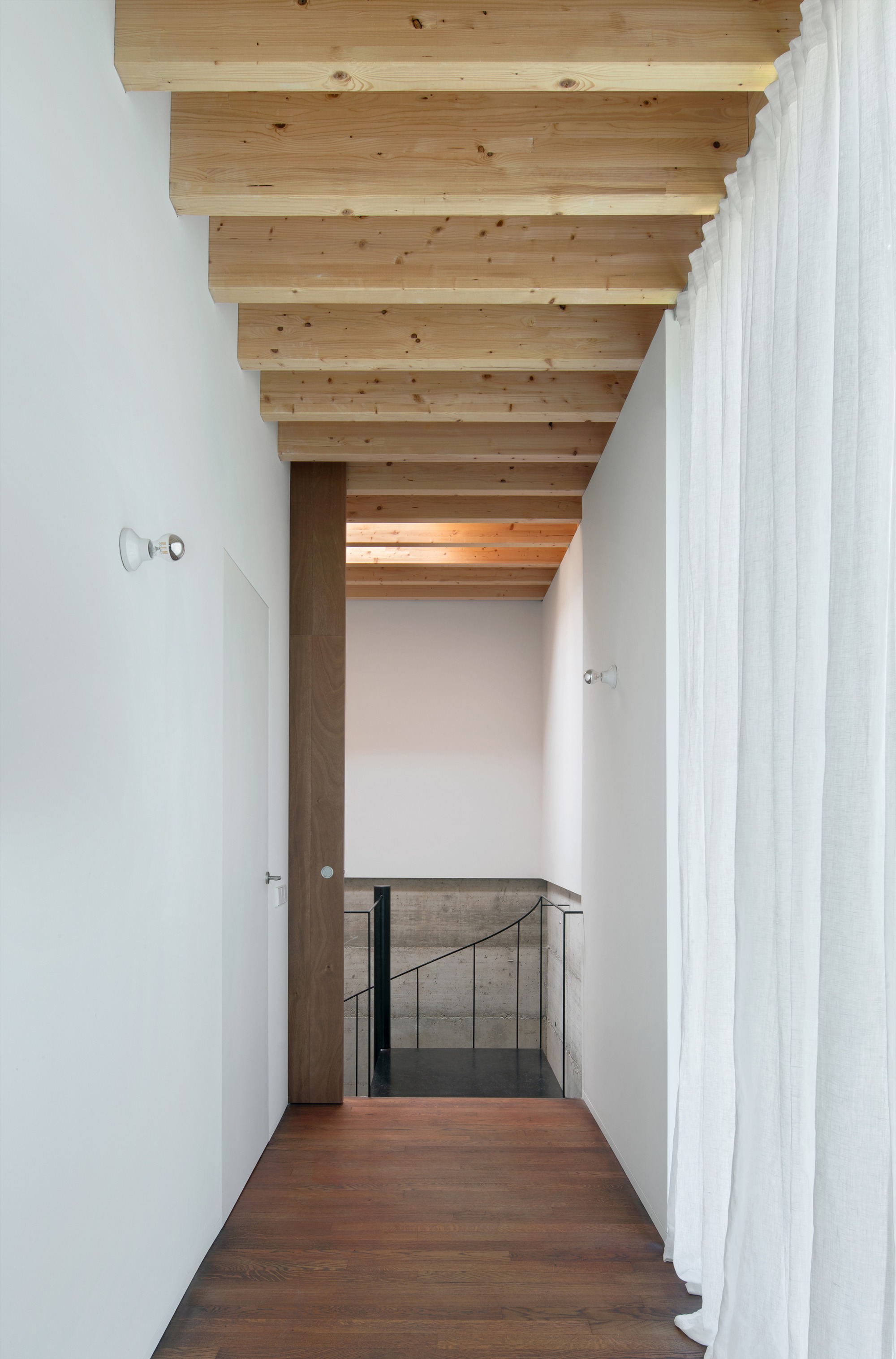 Corridor with exposed timber ceiling beams leading to glass-railed stairwell with white walls and timber flooring