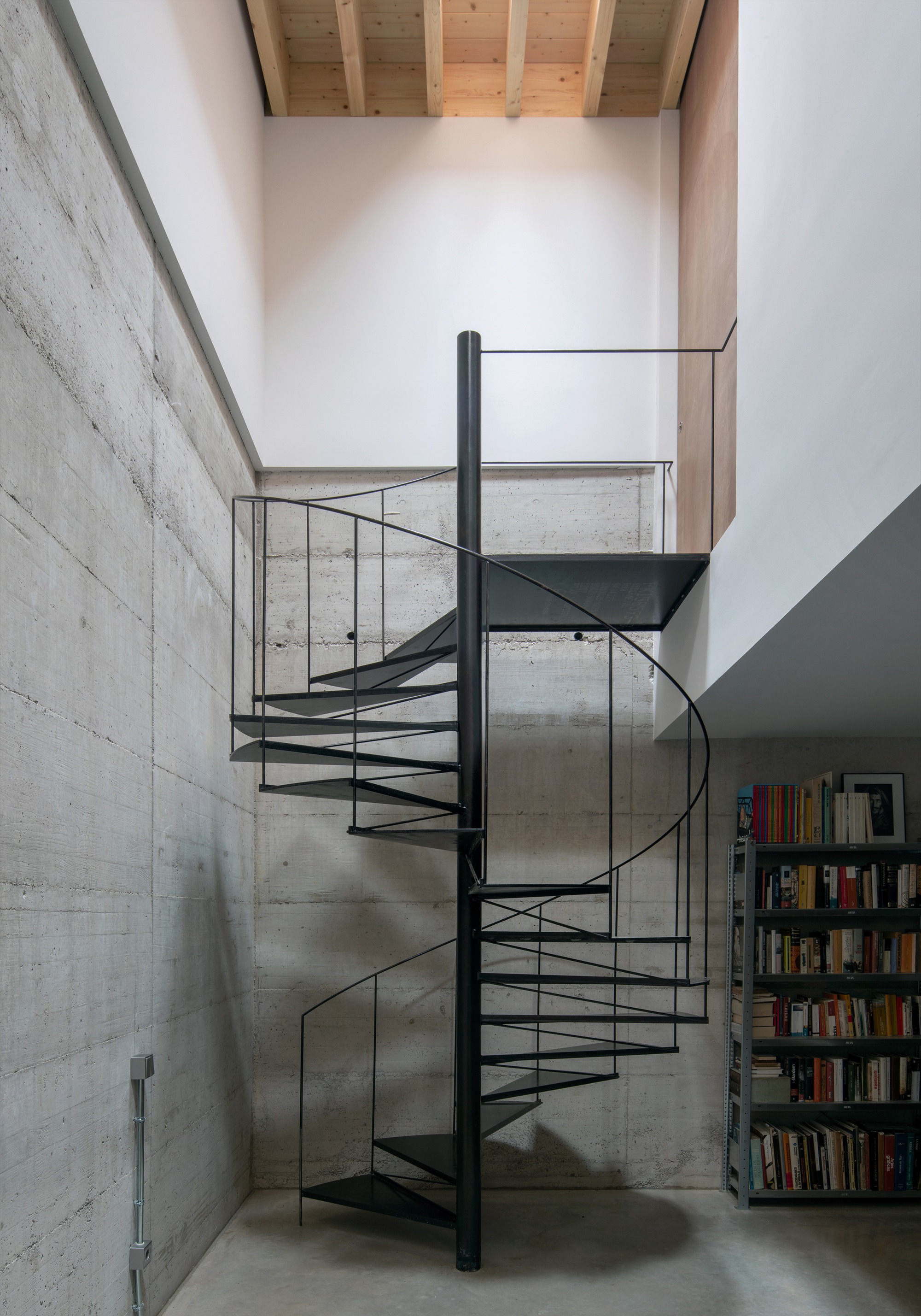 Black steel spiral staircase with glass railing against board-formed concrete wall with bookshelves visible below