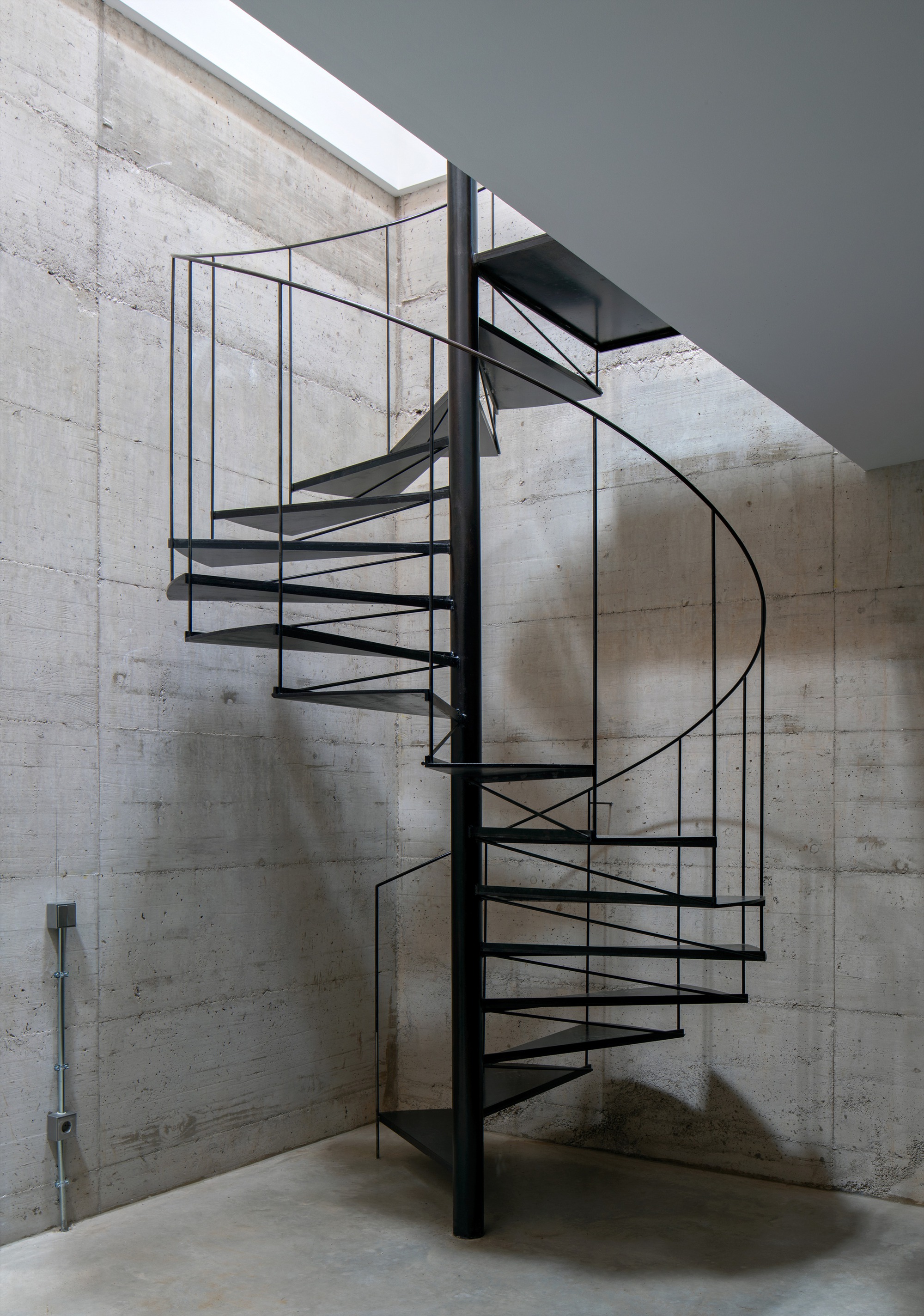 Spiral staircase rising toward skylight with board-formed concrete walls and natural light from above