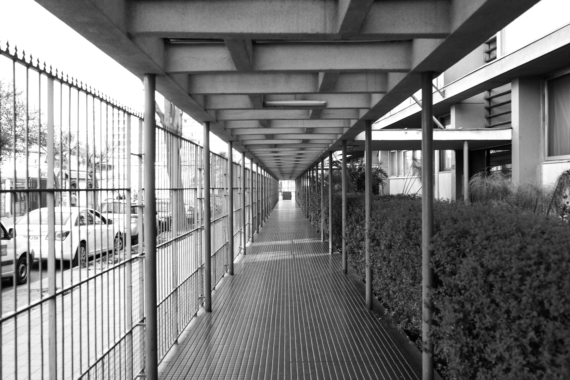 Long covered walkway with coffered concrete ceiling, metal fence and hedge along parked cars