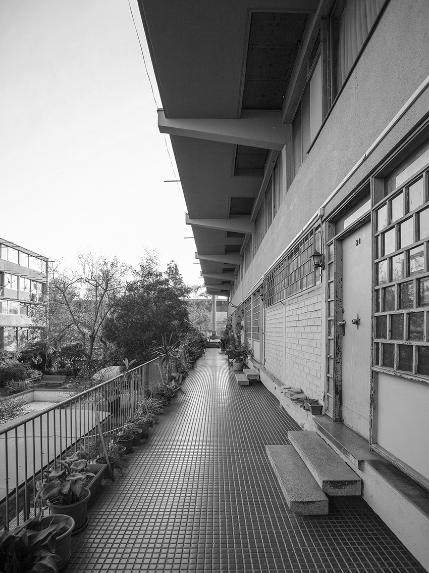 Covered external walkway with metal grating floor alongside planted beds and a metal railing