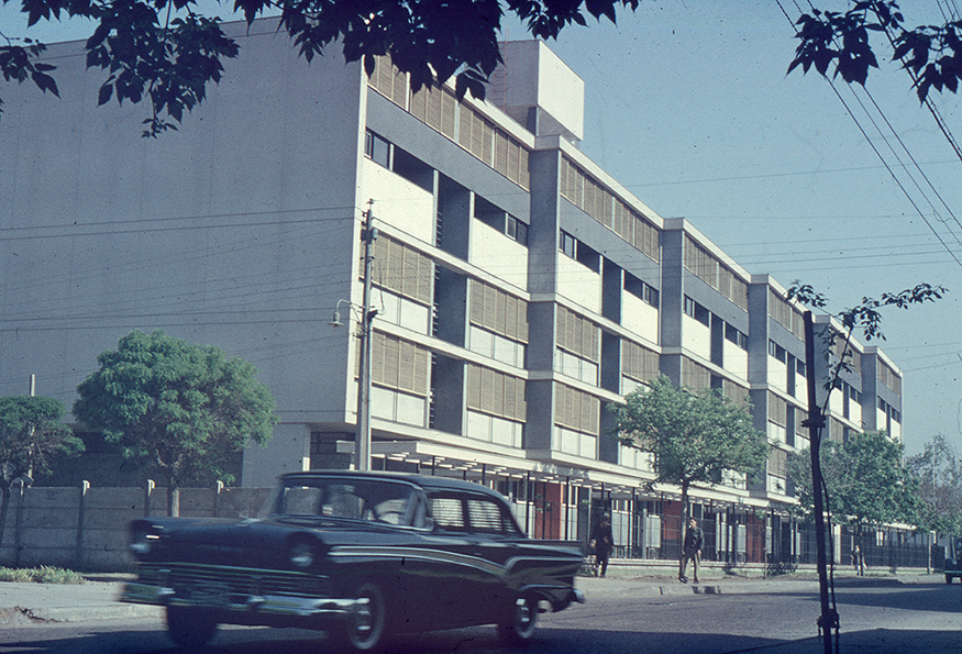 Street view of the gridded facade with alternating dark and light panels framed by foliage and a passing car