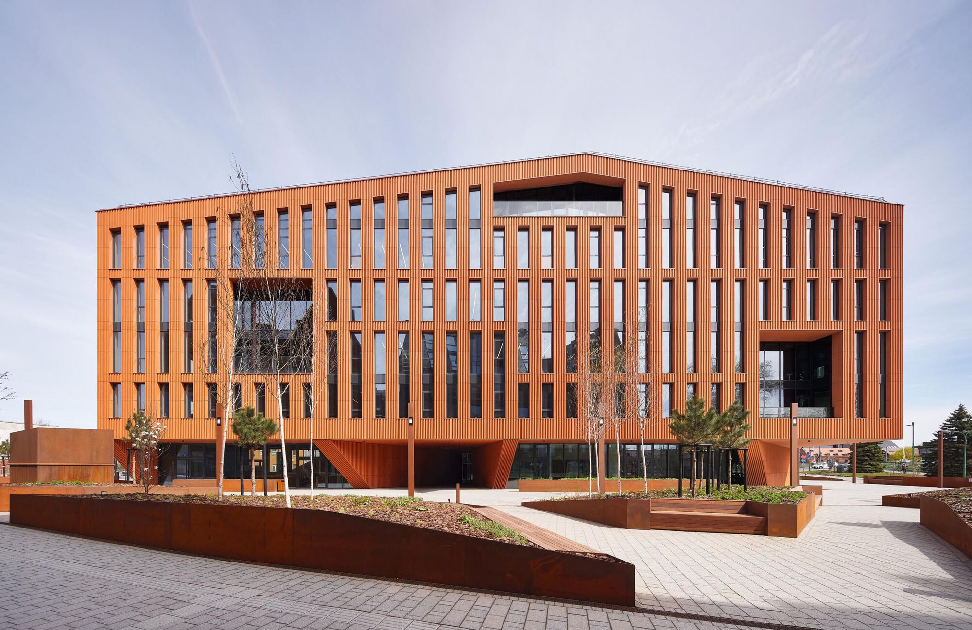 Plaza view of the terracotta facade with vertical slot windows and elevated volume above planted beds