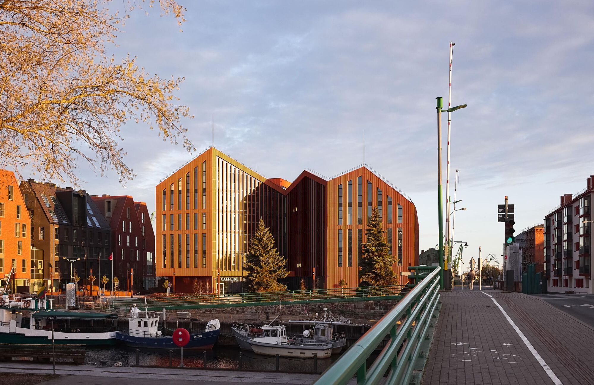 Cluster of gabled brick buildings with vertical window slots viewed across a canal at sunset