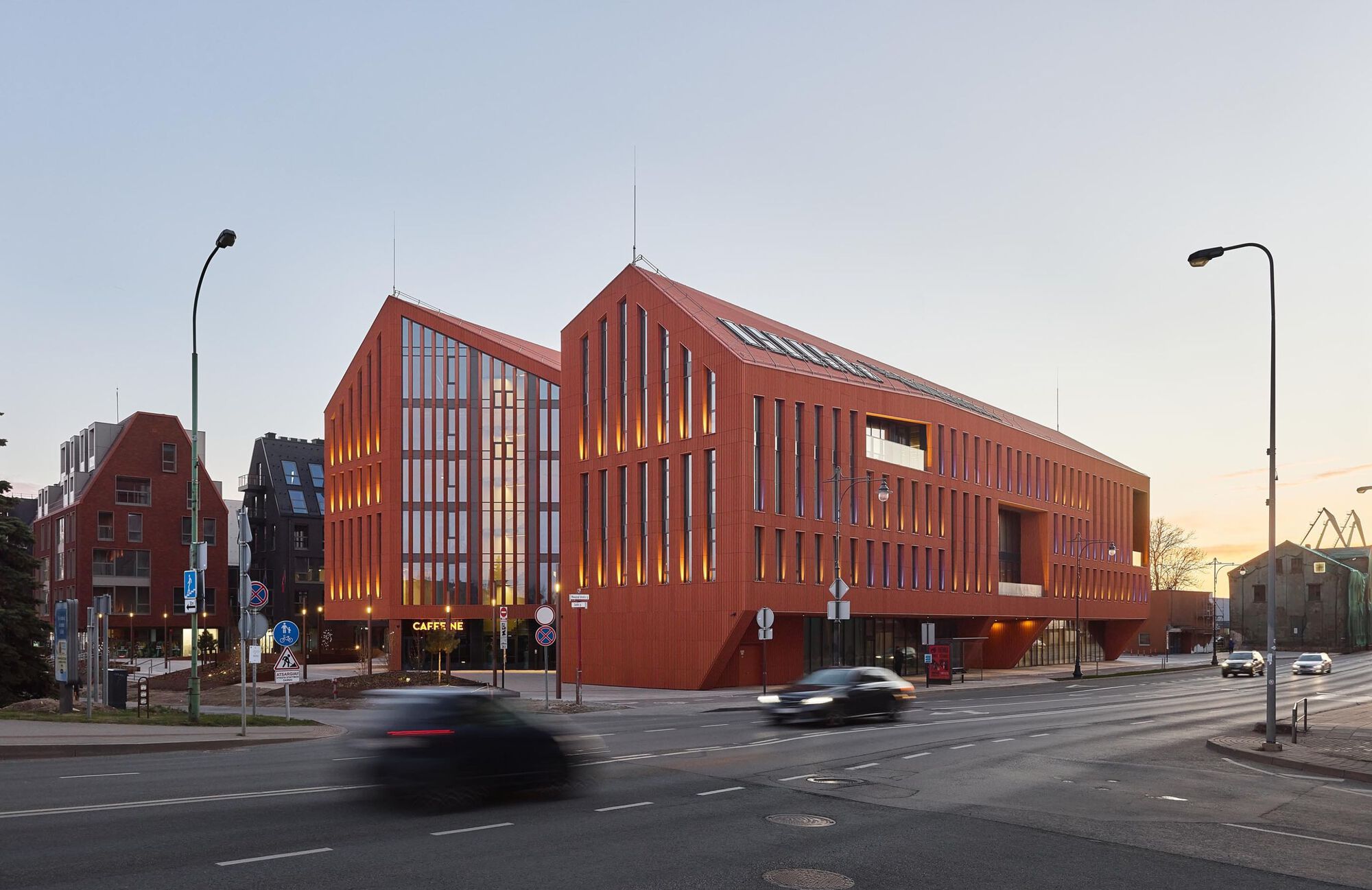 Street corner view of the red brick facade with vertical fenestration at dusk with passing cars