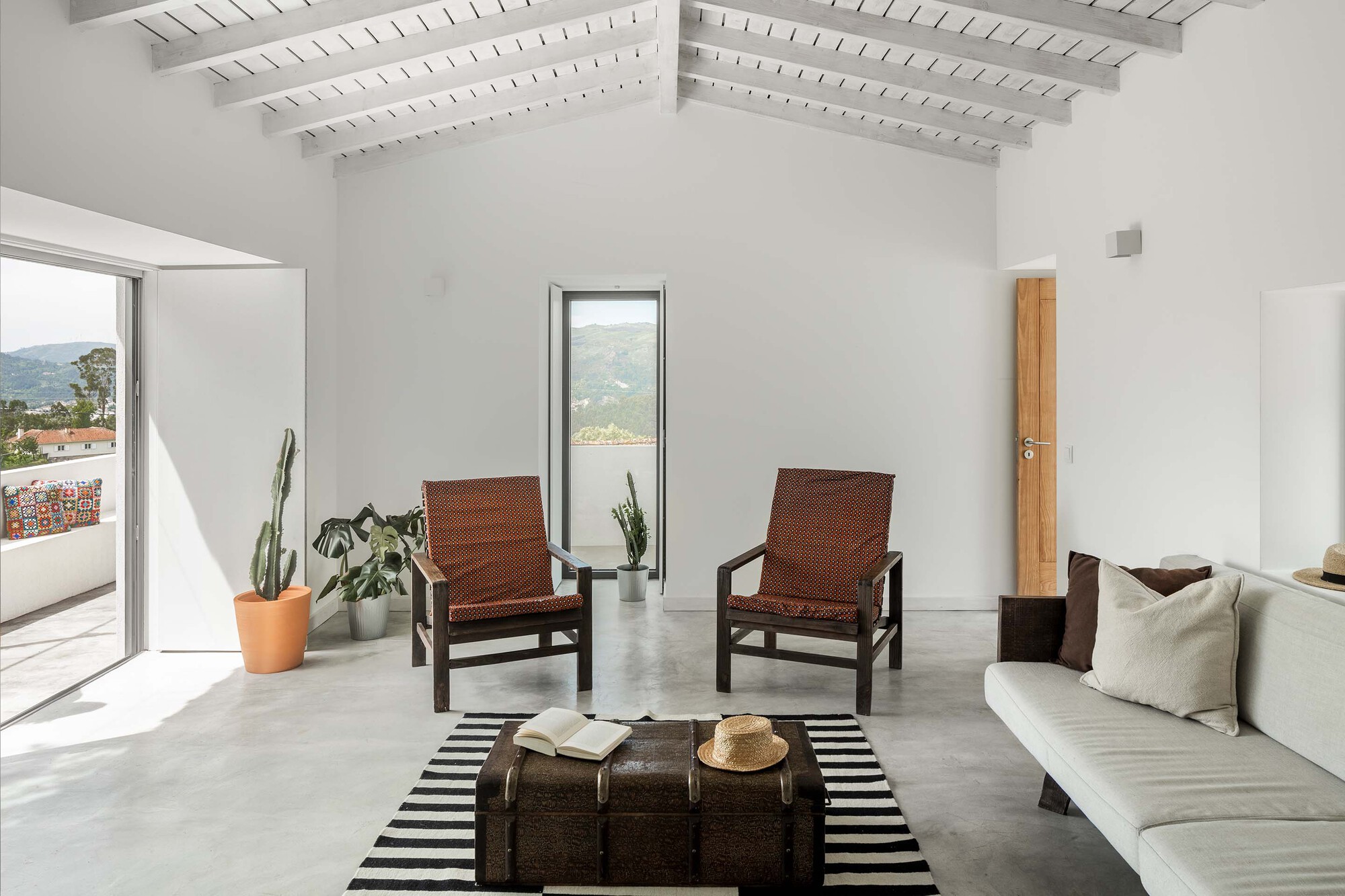 White living room with exposed timber ceiling joists and two woven chairs facing tall vertical windows