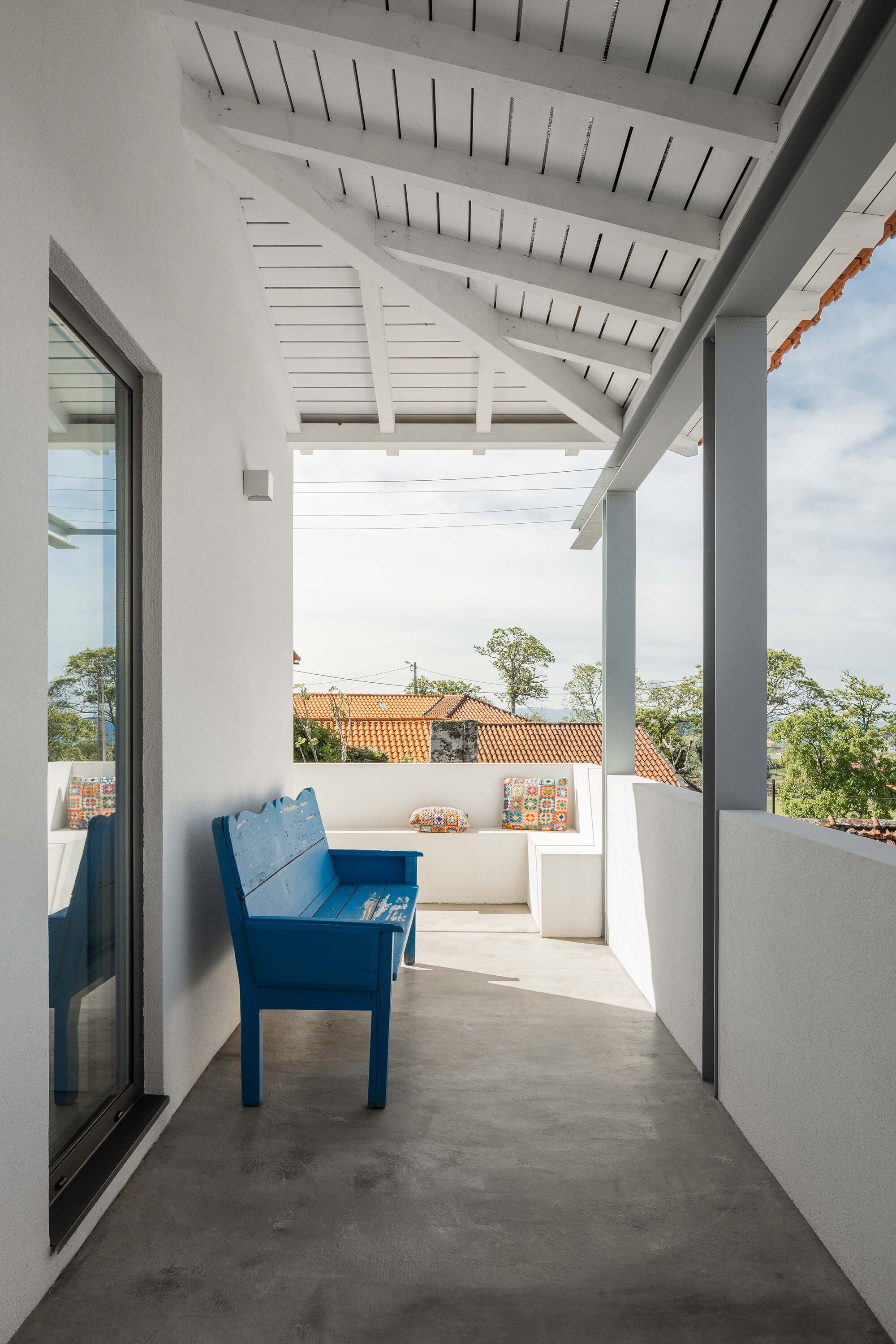 Covered terrace with painted timber ceiling, exposed rafters, and a blue bench overlooking clay-tiled rooftops
