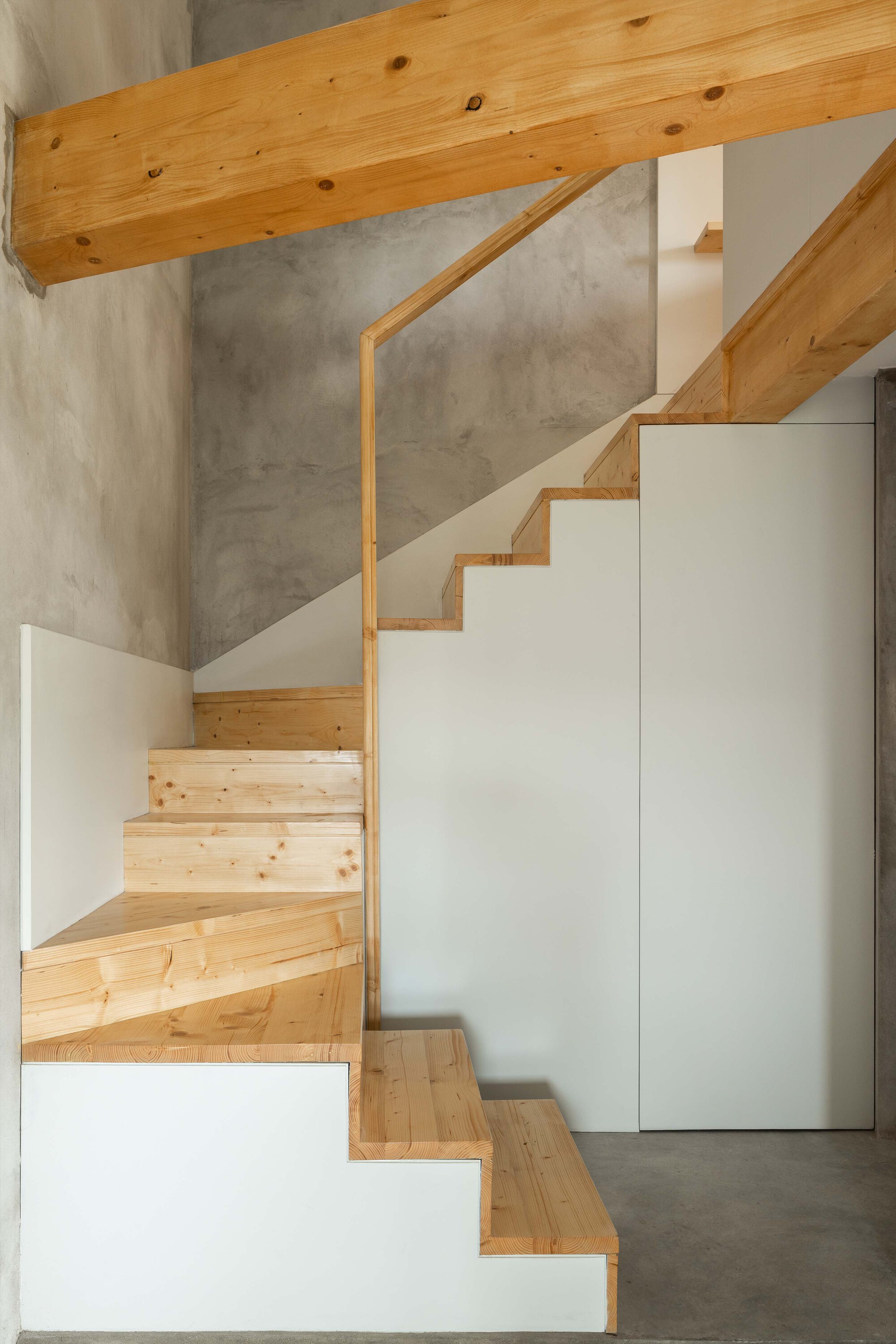 Timber staircase with white storage volumes beneath the treads against a concrete wall and exposed beam