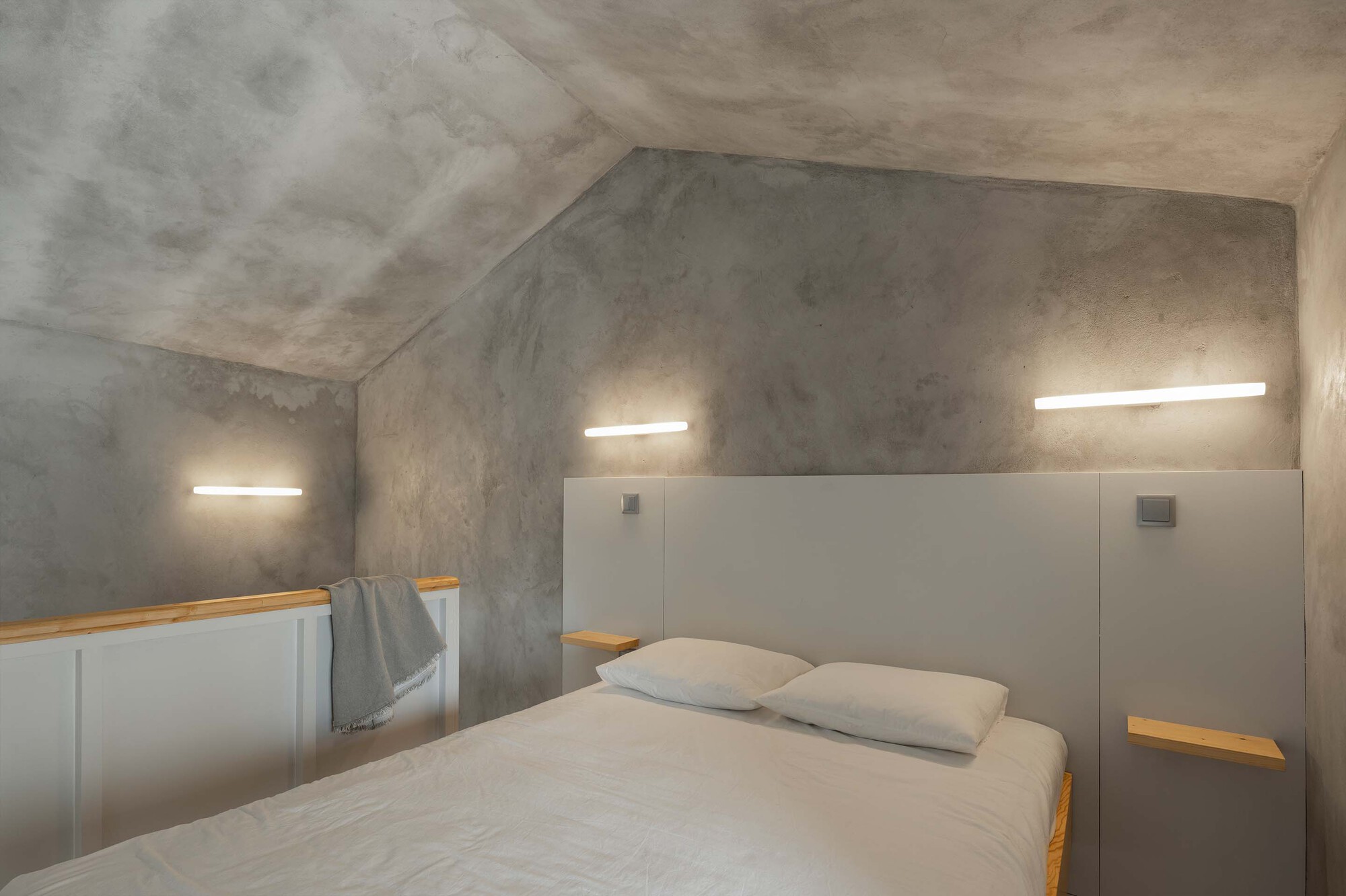 Bedroom with sloped concrete ceiling, horizontal recessed lighting, and grey paneled headboard with timber shelving