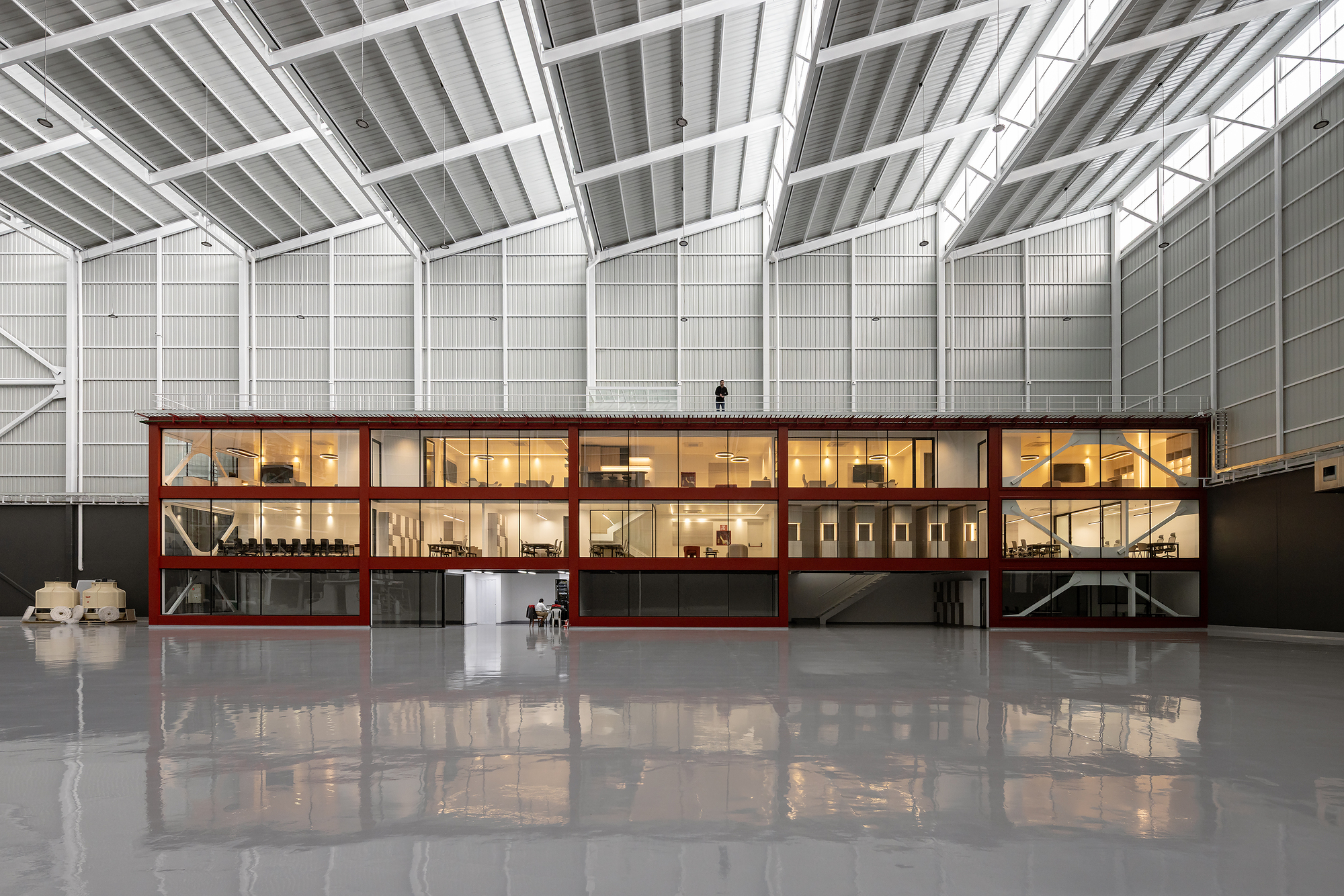 Two-story red steel-framed volume inserted within a vast white hangar with exposed roof trusses