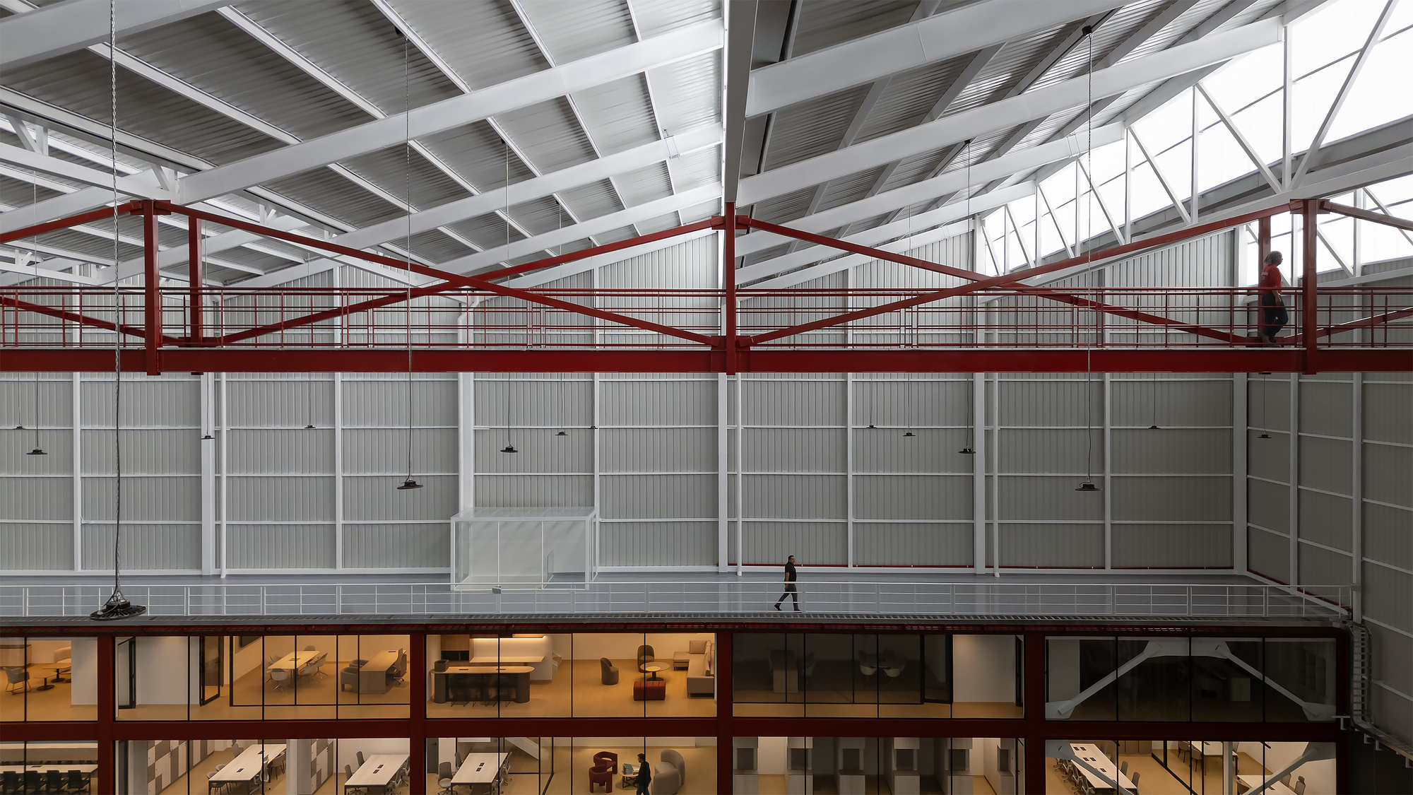 Red steel pedestrian bridge crossing the hangar interior beneath white corrugated metal walls with a person walking