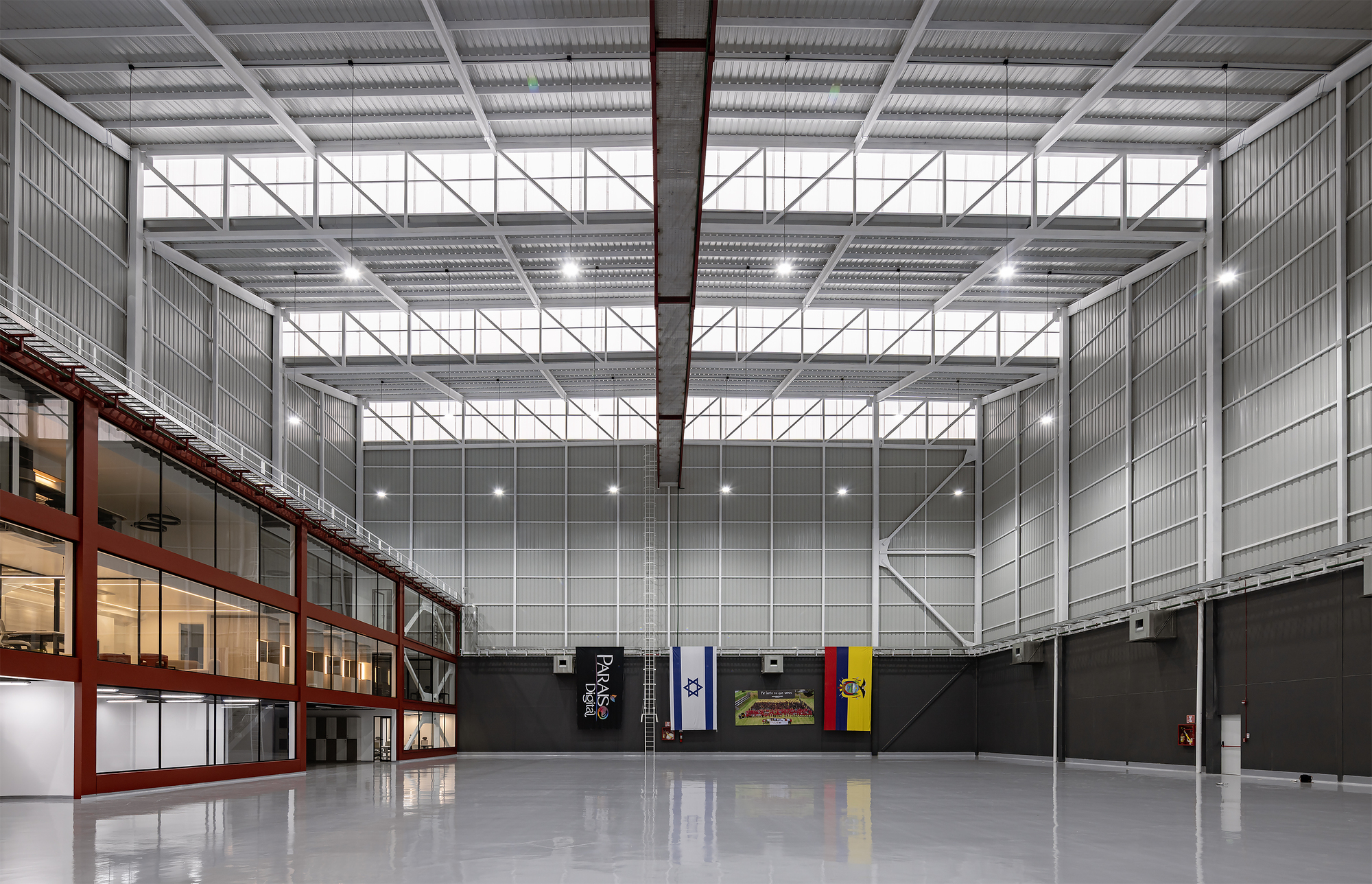 Interior view of a double-height hall with corrugated metal walls, exposed truss ceiling, and two-story red-framed windows