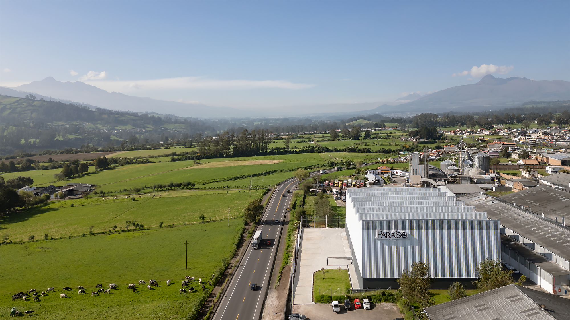 Aerial view of the corrugated metal-clad building beside a highway with green fields and mountains beyond