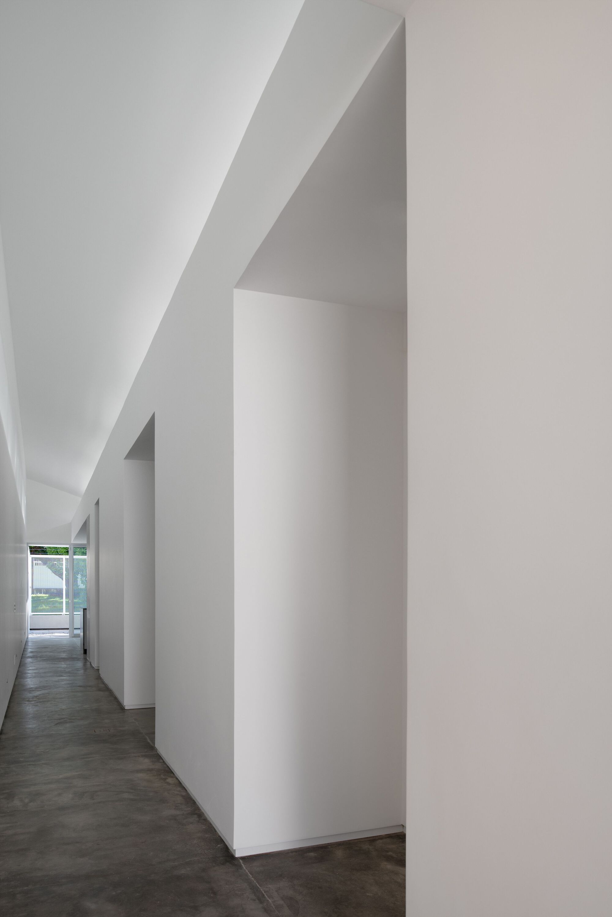 Narrow corridor with angular white walls and sloped ceiling leading toward a distant window