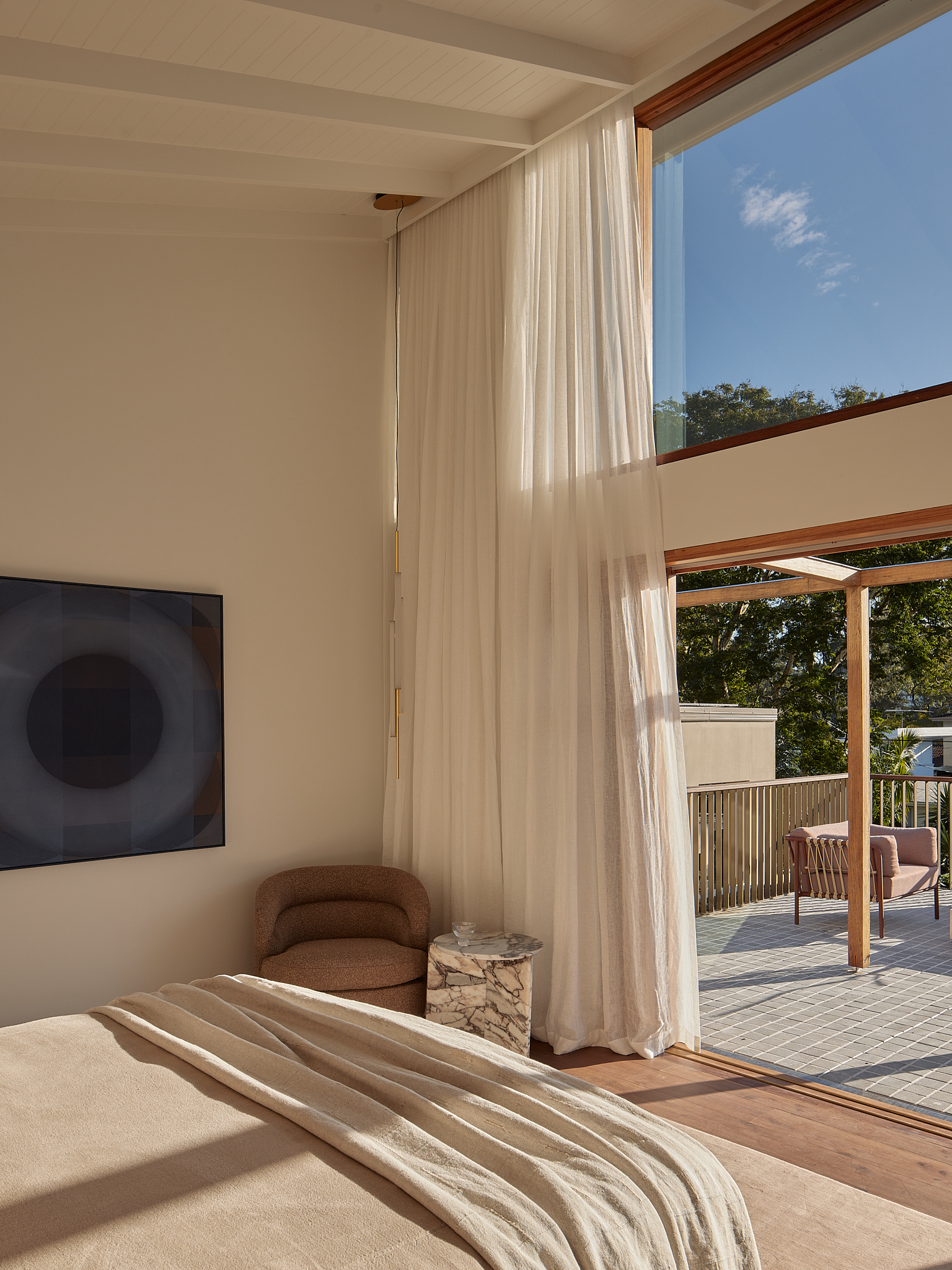Bedroom opening to timber-framed terrace through sliding glass doors with flowing white curtains in daylight