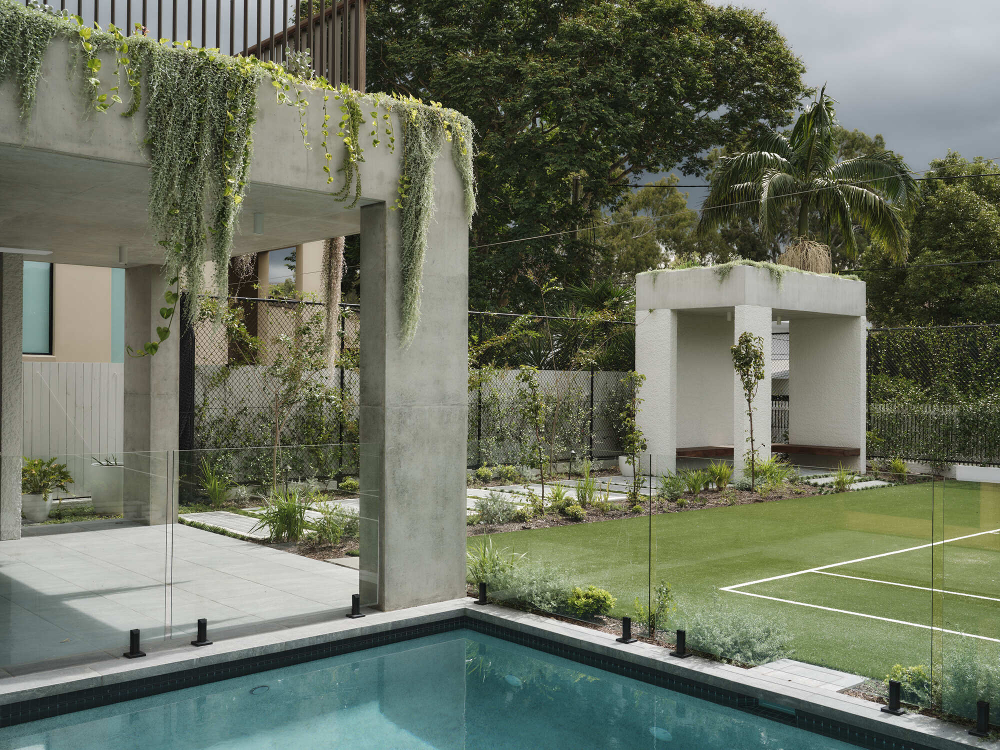 Rear garden view showing concrete pergola with trailing vines, glass balustrade pool edge and lawn under cloudy skies
