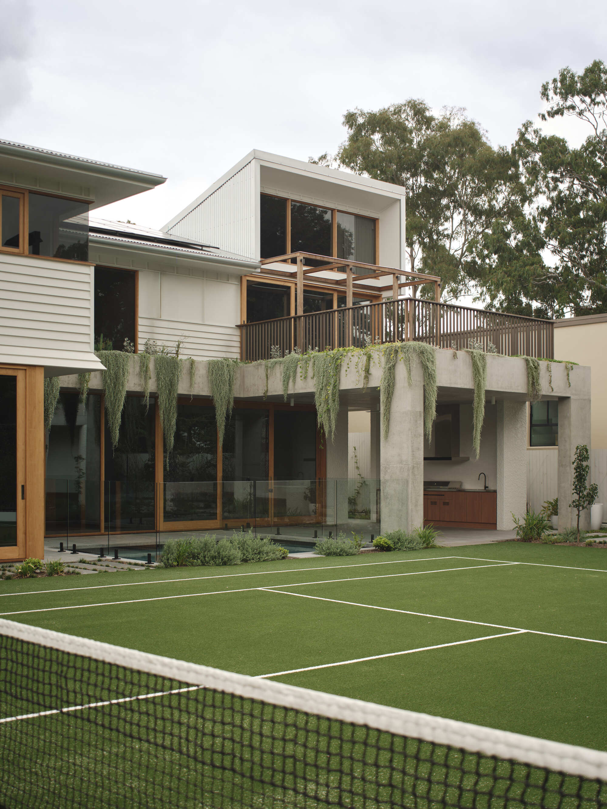 Garden facade with concrete planter wall, cascading greenery, timber balcony and tennis court under overcast sky