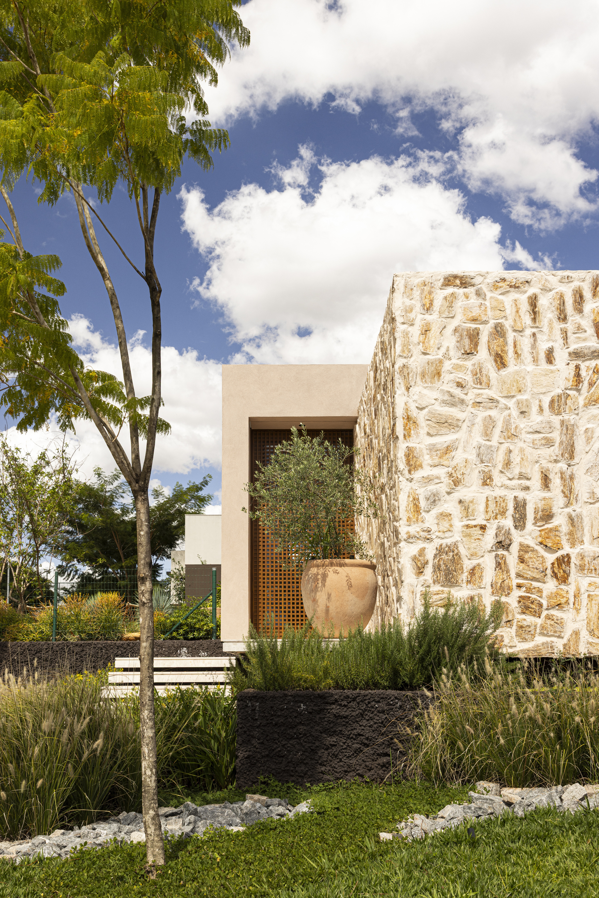 Entry facade with rough limestone wall, perforated timber screen, and planted garden beds